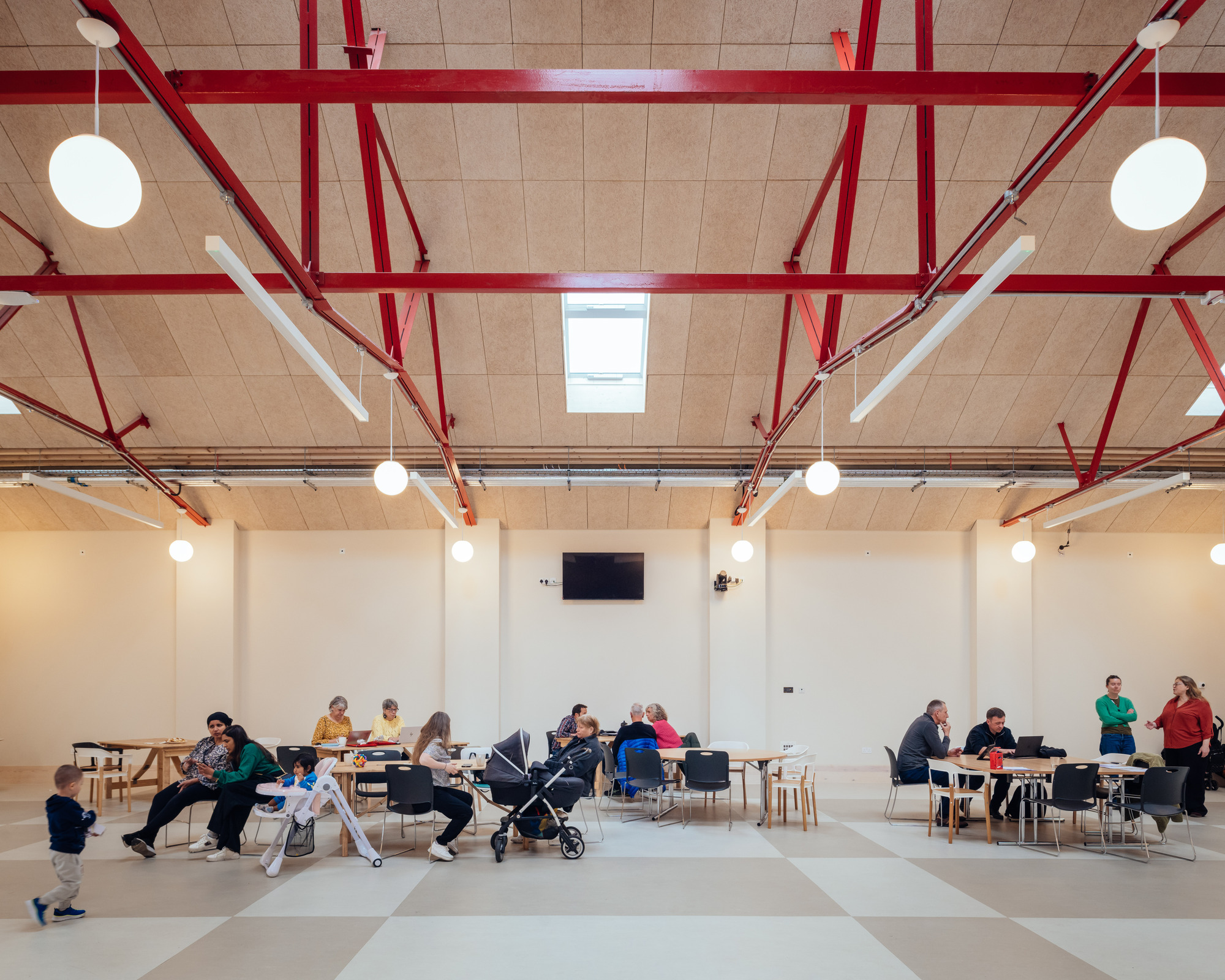 Wide view of the hall with families at tables, a pushchair and a toddler walking beneath red trusses and globe lights