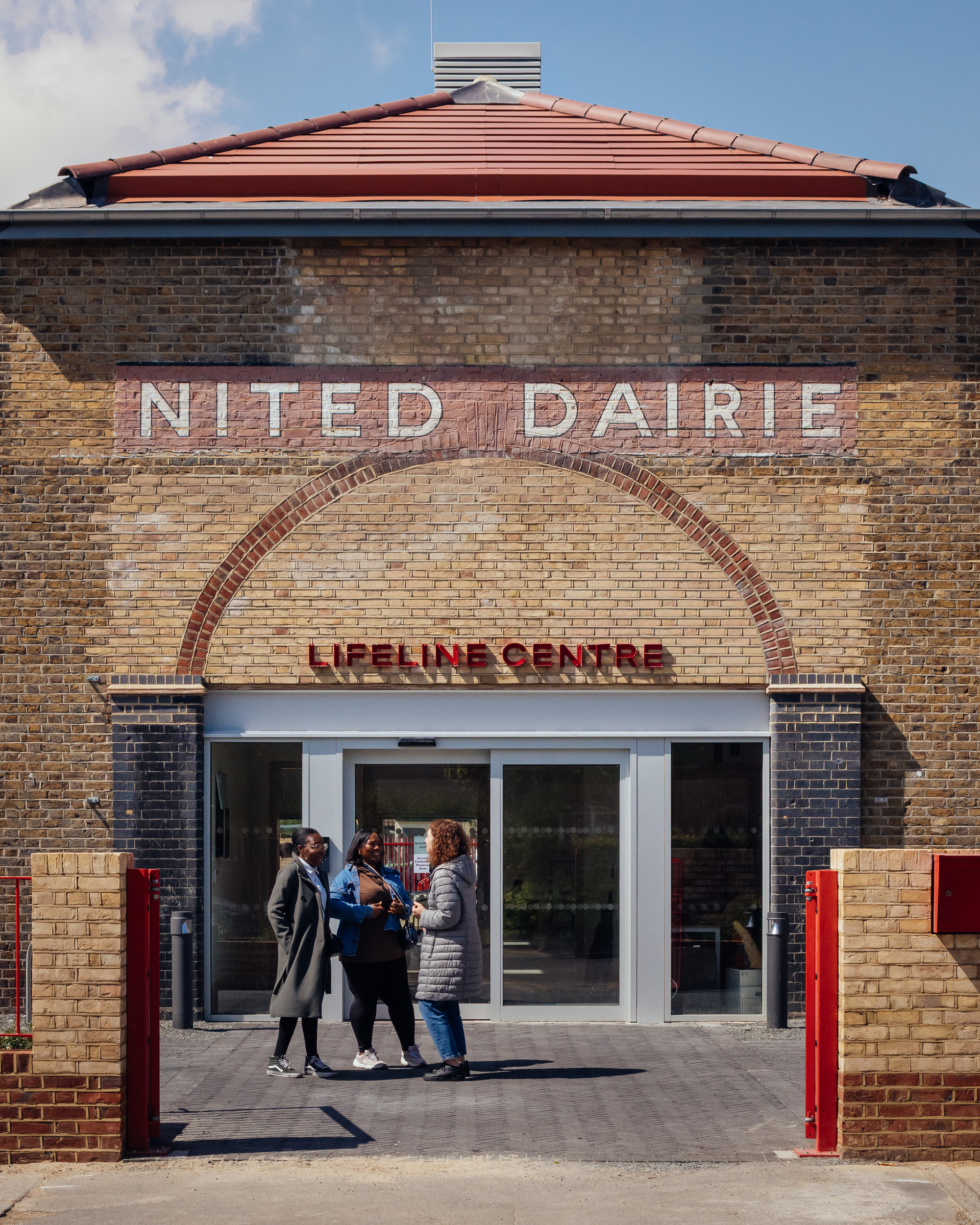 Close view of the brick entrance with red brick arch and painted sign, three people chatting in front of sliding glass doors