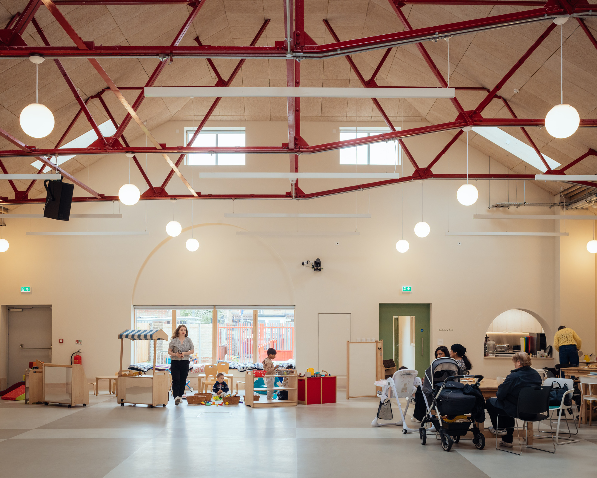 Double-height hall with red steel trusses, globe pendants and timber ceiling panels, with parents and toddlers gathered at play tables below