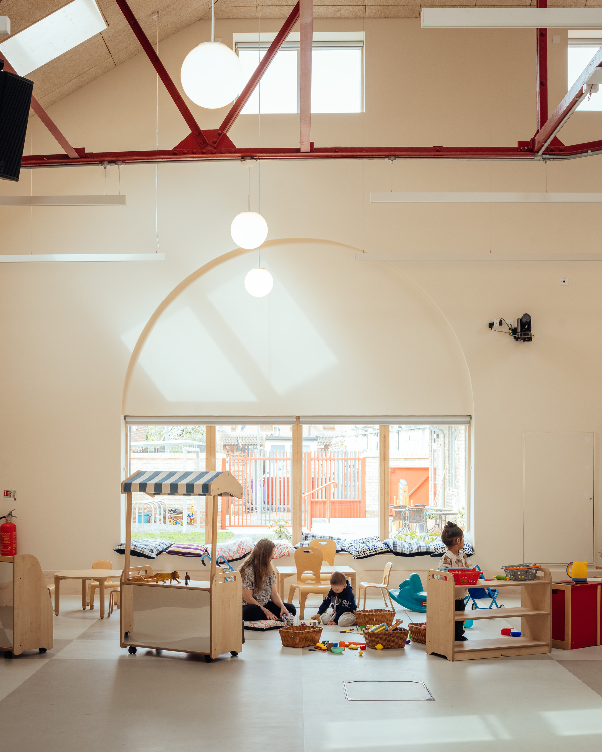 Arched window above a low cushioned bench, with children playing among wooden shelves and a toy stall in soft daylight