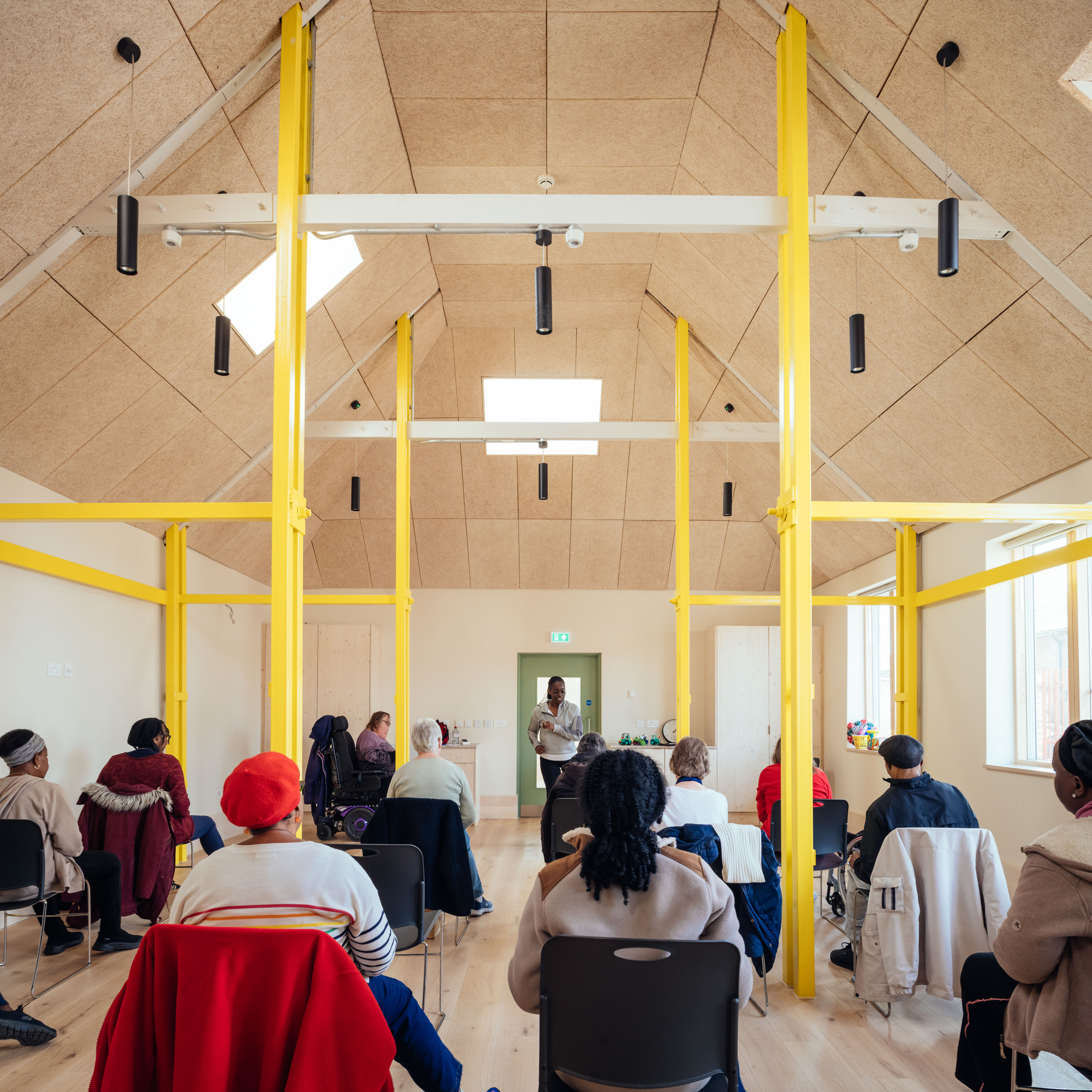Pitched timber-panel ceiling framed by yellow steel columns, with seated adults facing a speaker near a green door