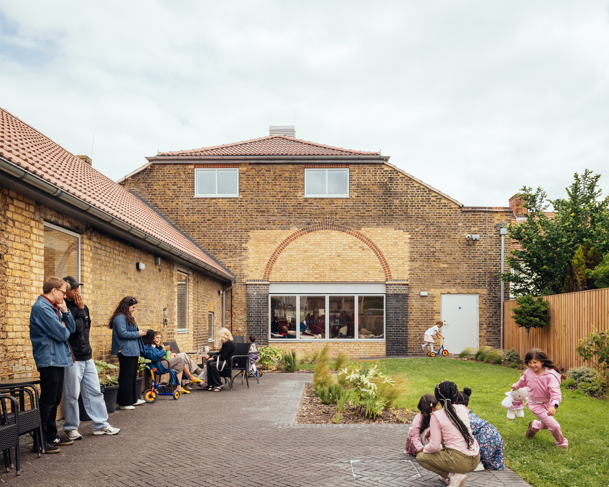 Rear courtyard of yellow brick walls with an arched window, where children play on grass and adults chat on the paving
