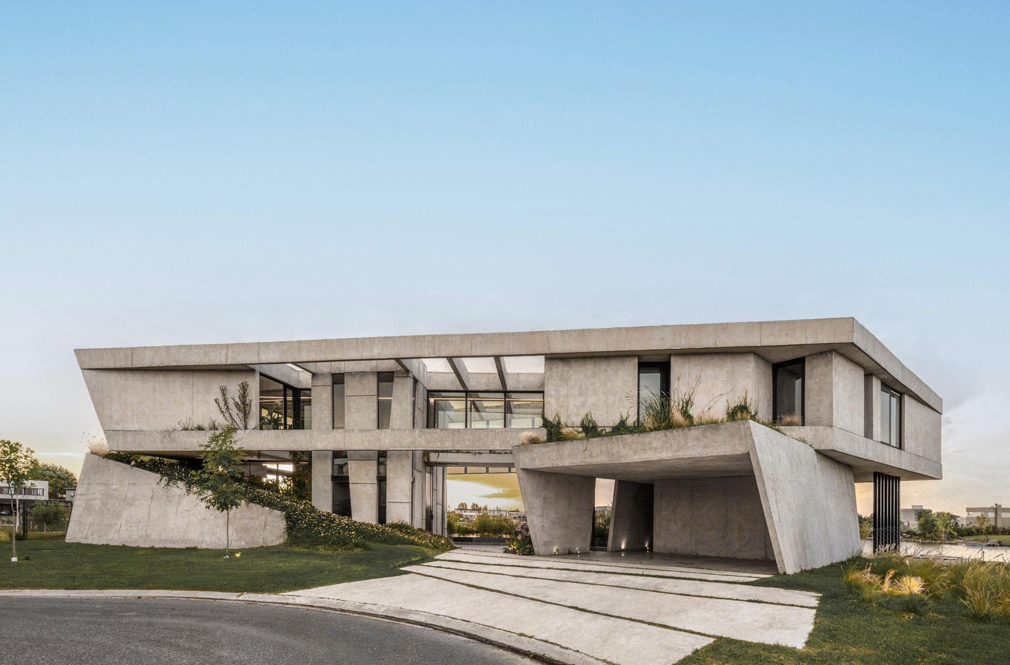 Corner view of the board-formed concrete house with cantilevered upper volume and angled carport wall at dusk, planted terrace along the roofline