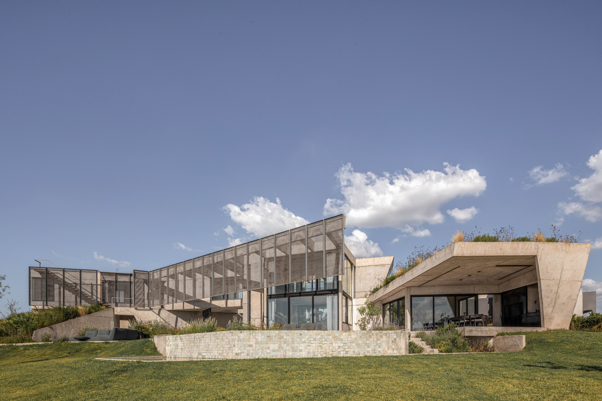 Concrete house on a grassy rise with a perforated metal screen over glazing and a planted roof under scattered clouds