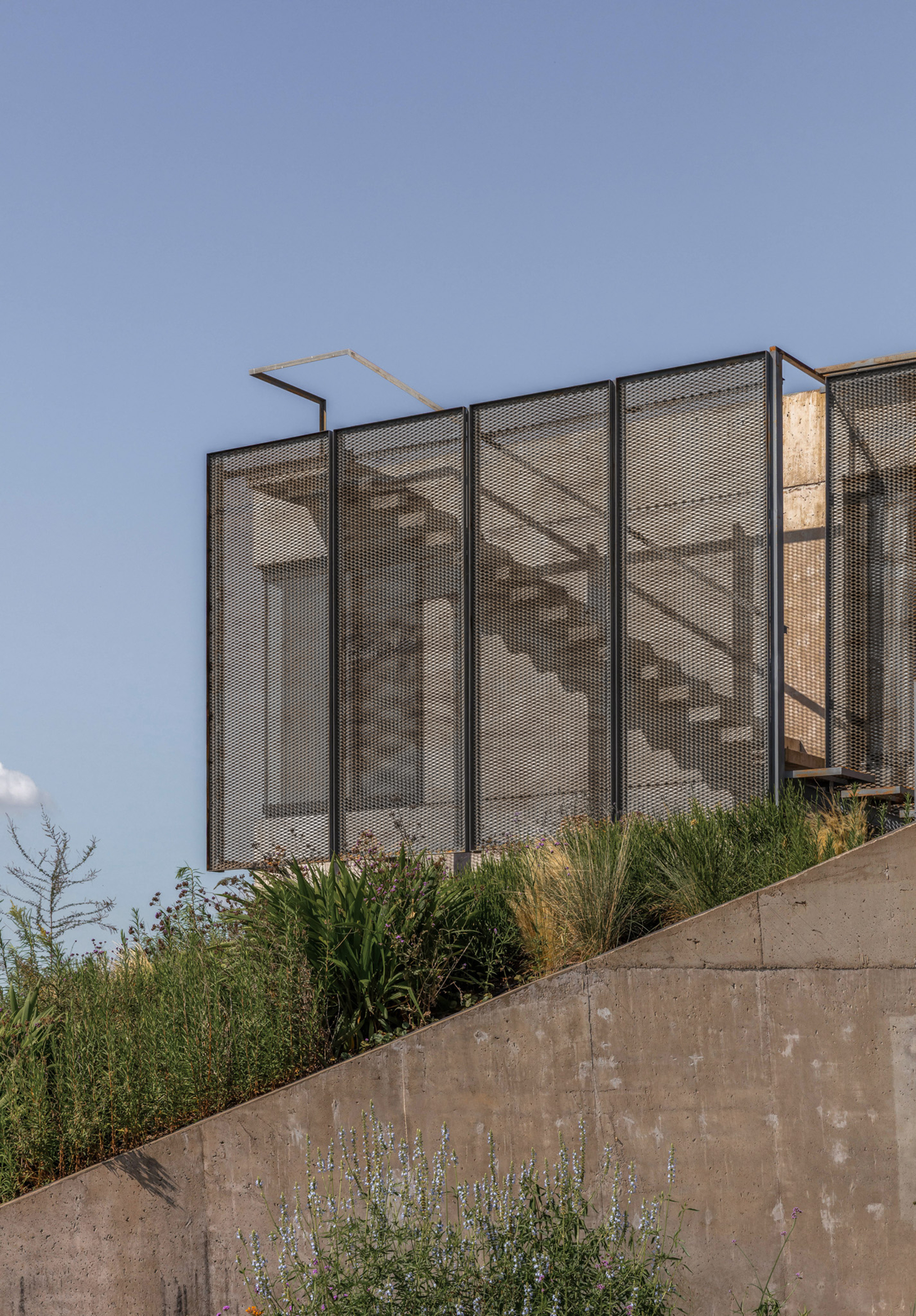 Perforated metal screen enclosing an exterior stair, set above a sloping concrete wall and tall grasses in clear sky