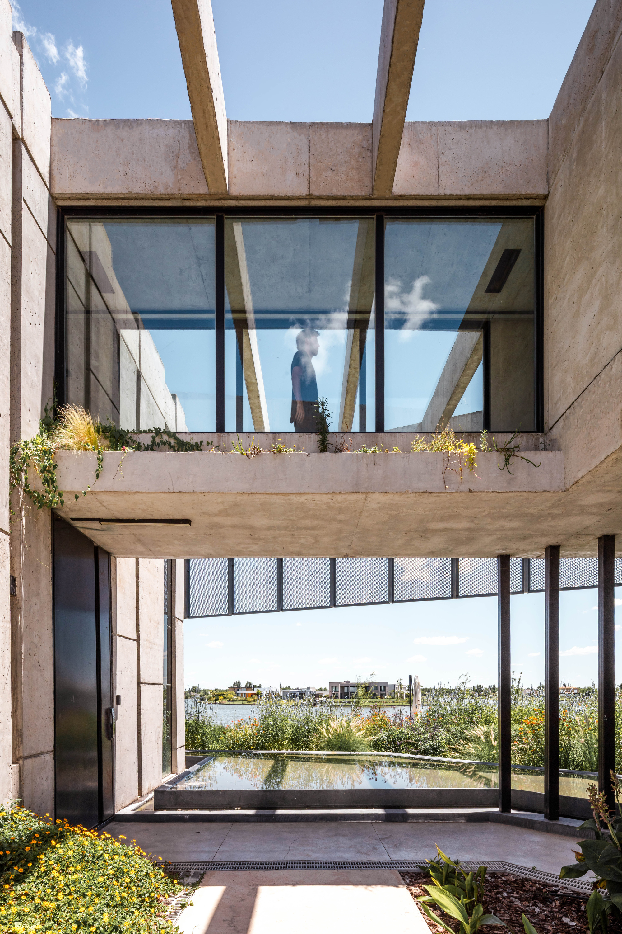 Entrance courtyard with a planted concrete bridge above a reflecting pool, a person visible through the upper glazing in daylight