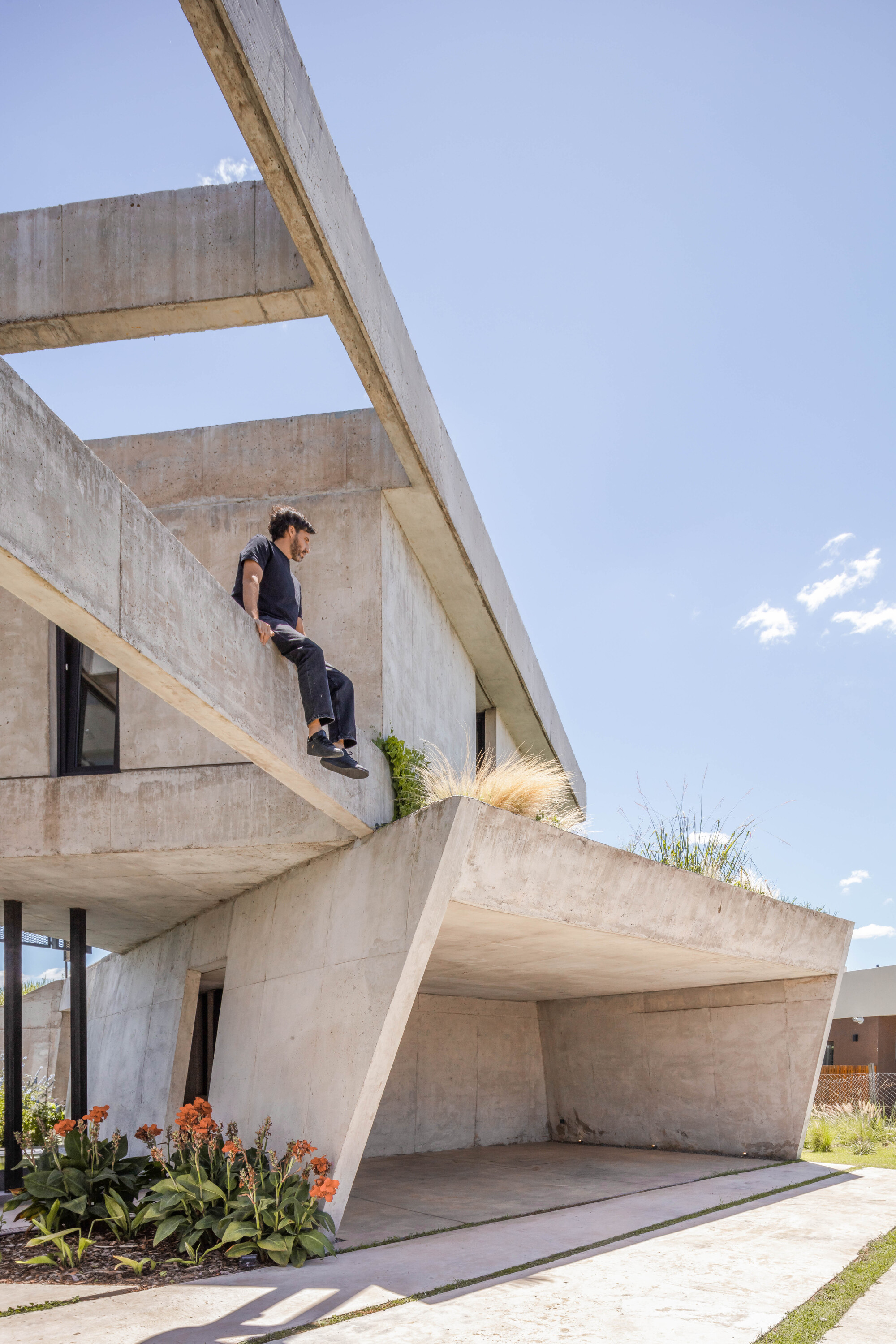 A man sits on the edge of a concrete beam above the angled carport wall, with orange flowers in the foreground
