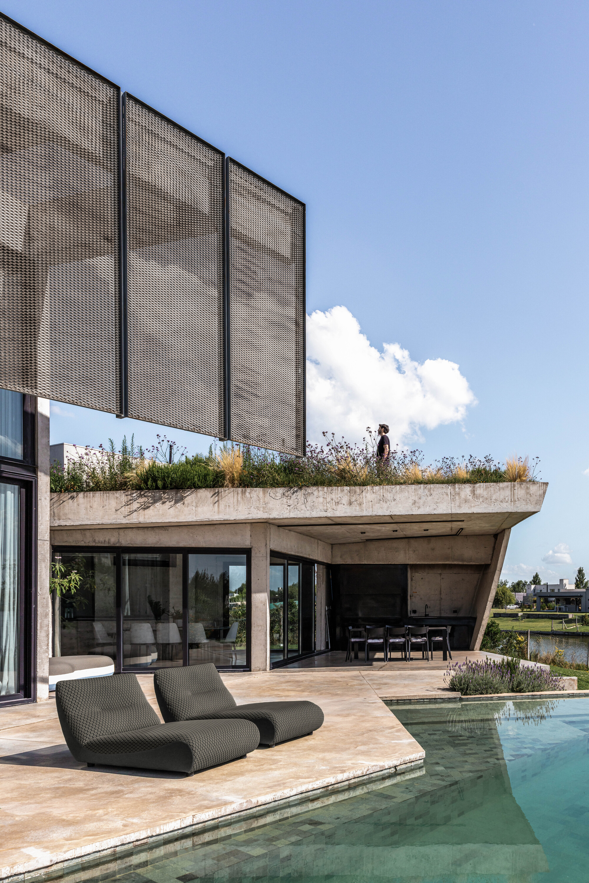 Perforated metal screen panels above a planted concrete terrace edge, with lounge chairs beside a turquoise pool under a blue sky