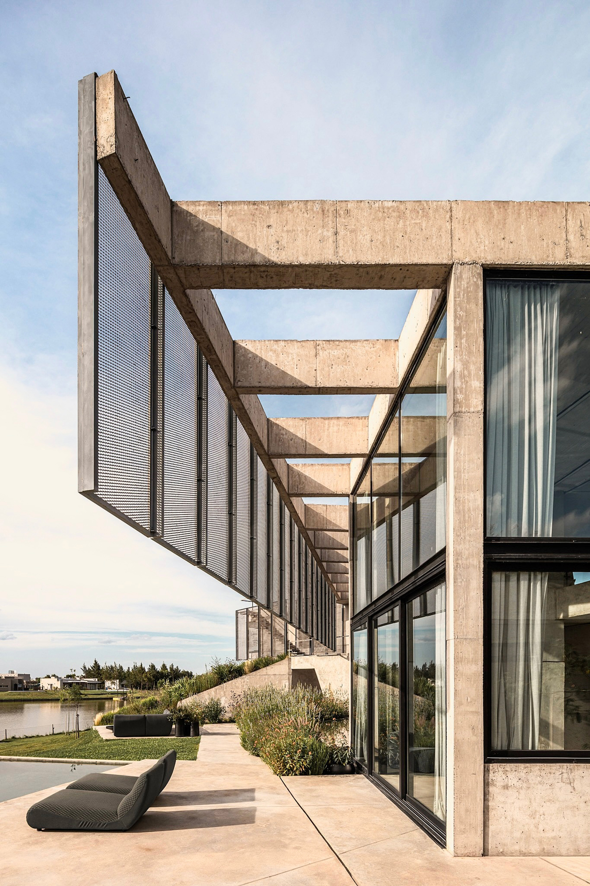 Receding concrete beams and mesh screen panels along a glass corner, with a lounge chair on the paving beside the lake