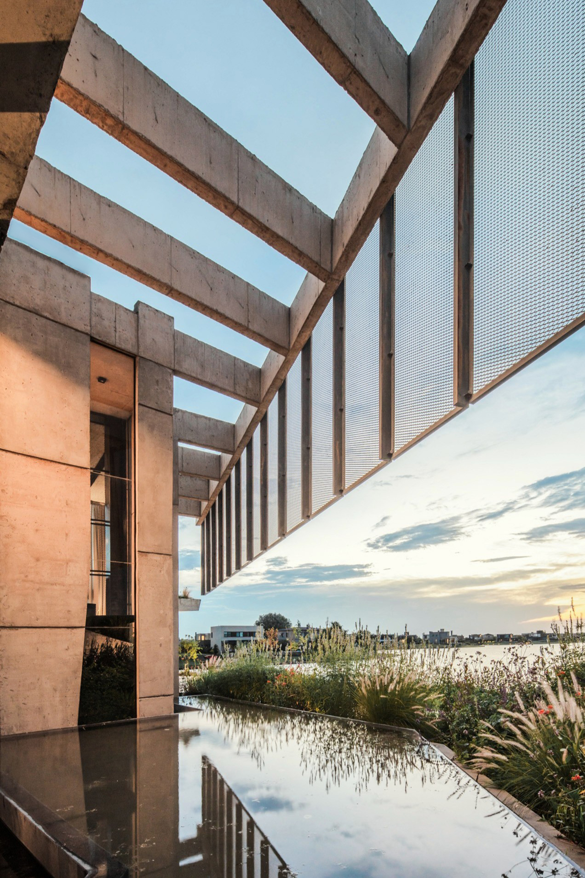 Concrete beams and slatted mesh screen seen from a reflecting pool, with grasses and a sunset sky over the lake