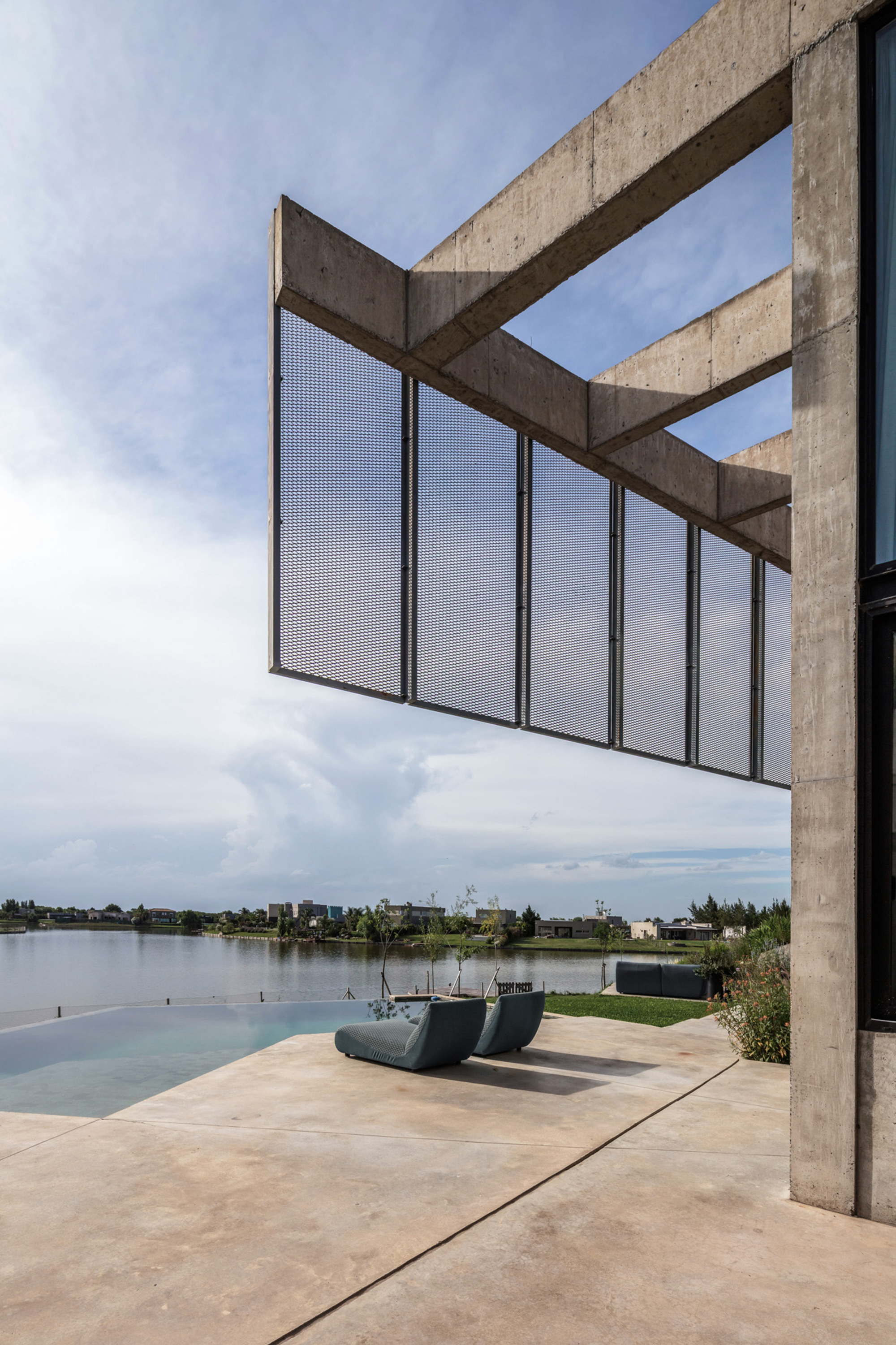 Cantilevered concrete frame with hanging perforated metal screen above a pool terrace and two lounge chairs facing a lake under cloudy sky