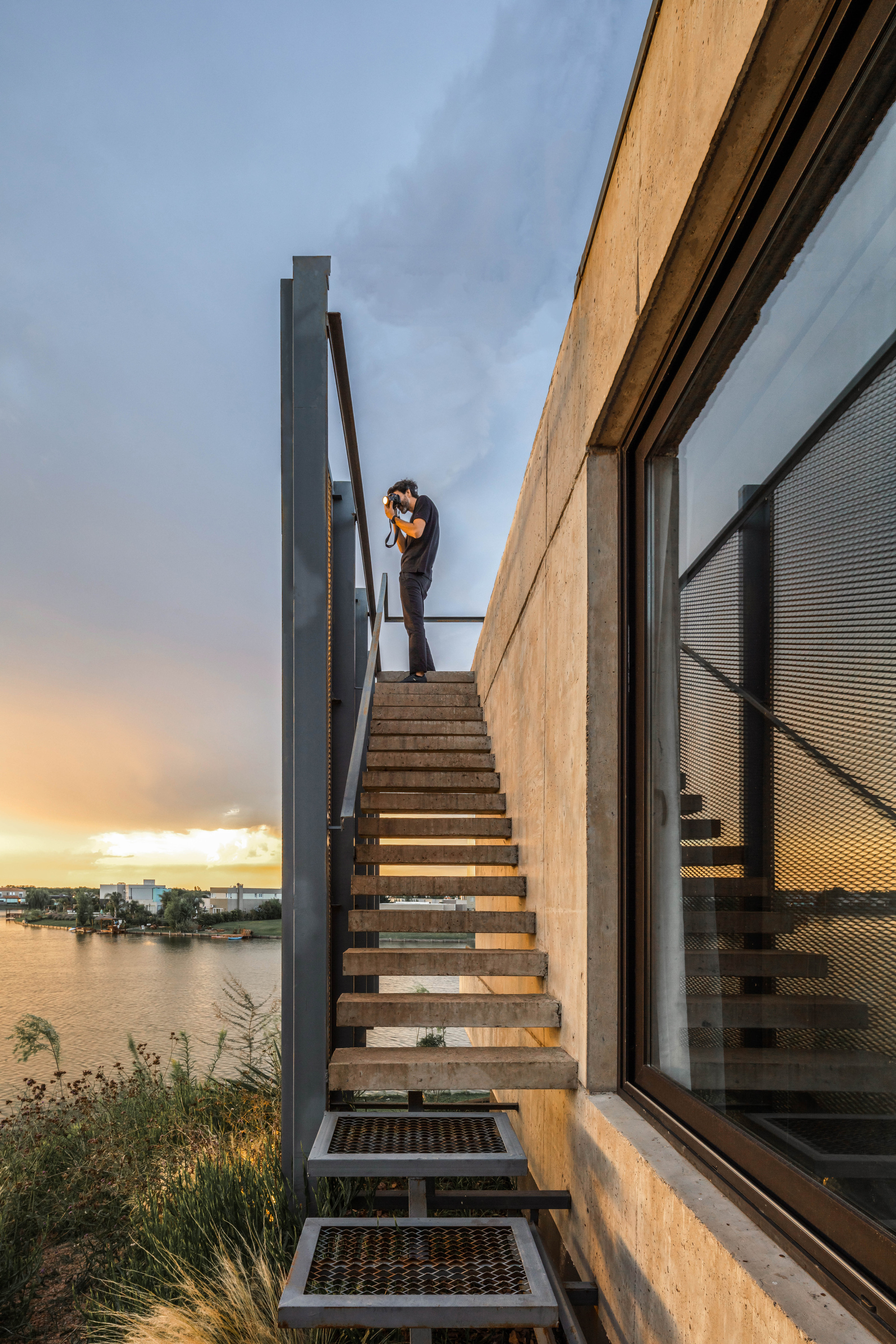 Photographer standing on a steel and timber exterior stair beside a board-formed concrete wall at sunset over the water