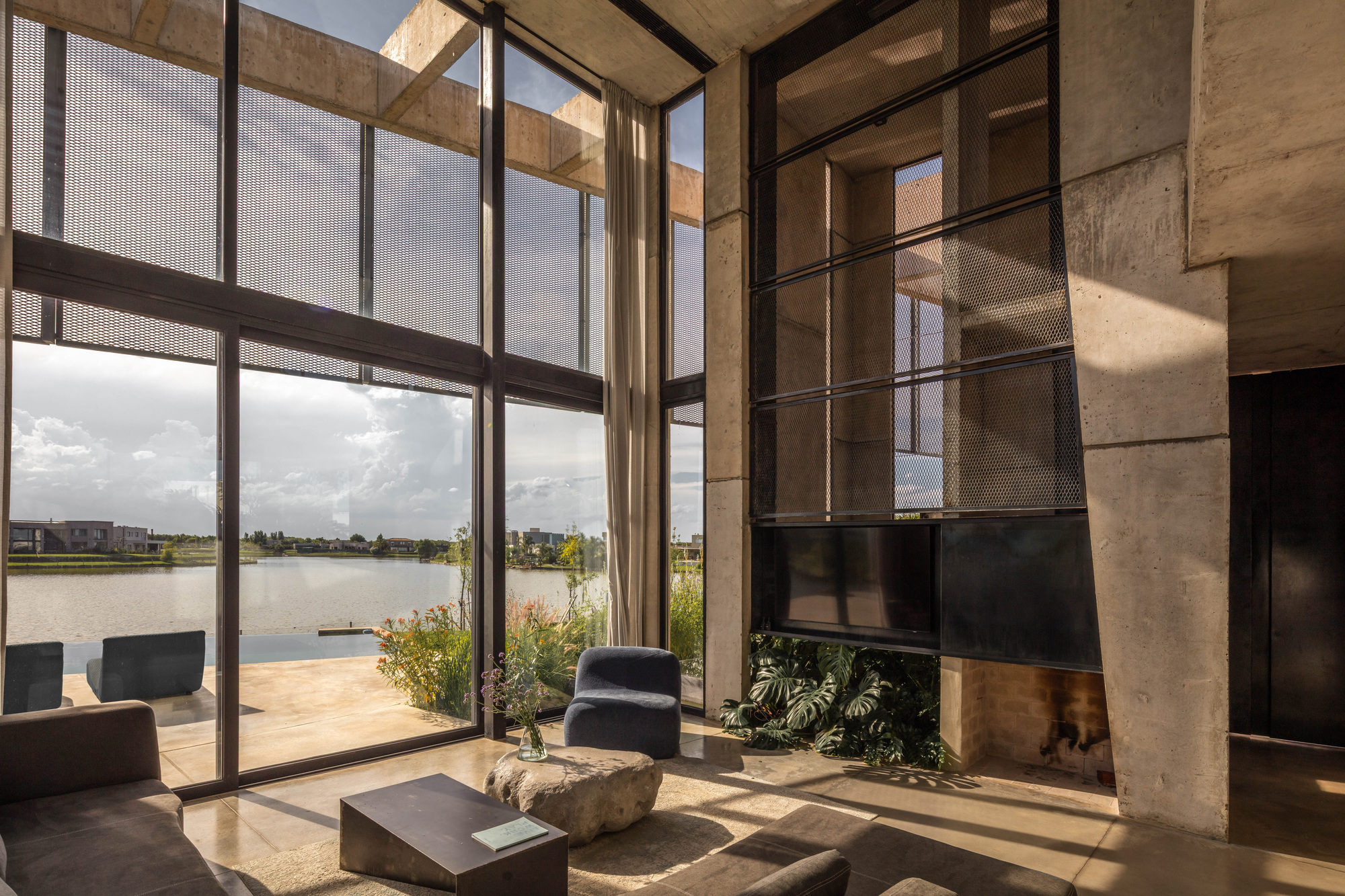 Double-height living room with floor-to-ceiling glazing, mesh screens and a concrete column, looking out over a pool and lake