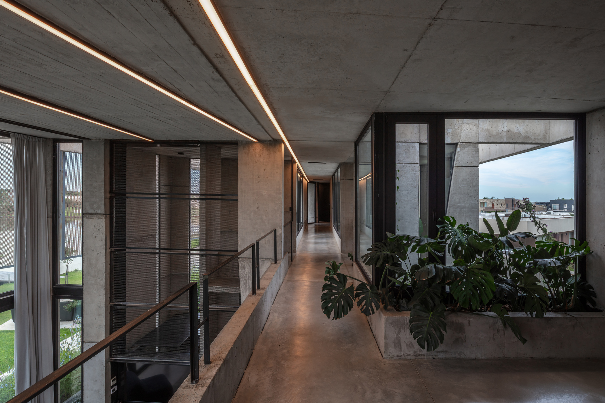 Concrete corridor with linear ceiling lights, steel railings and a monstera planter beside a glazed corner at dusk