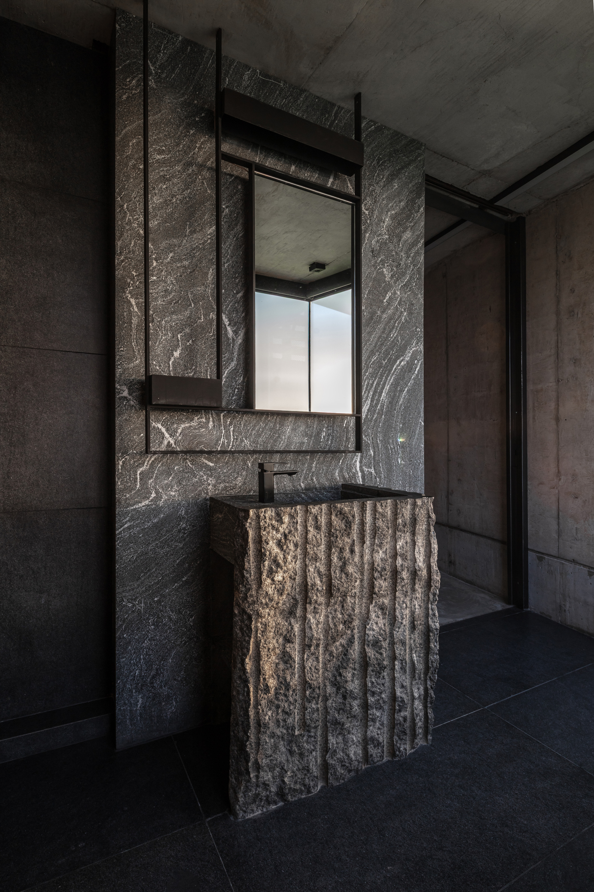 Rough-cut stone pedestal basin against a veined dark stone wall with a hanging mirror and black tap
