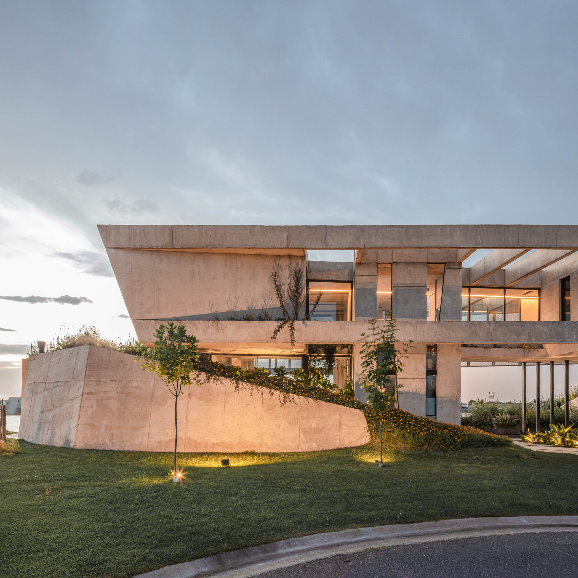 Exposed concrete volume with a curved retaining wall and ivy-covered slope on a lawn under a cloudy evening sky
