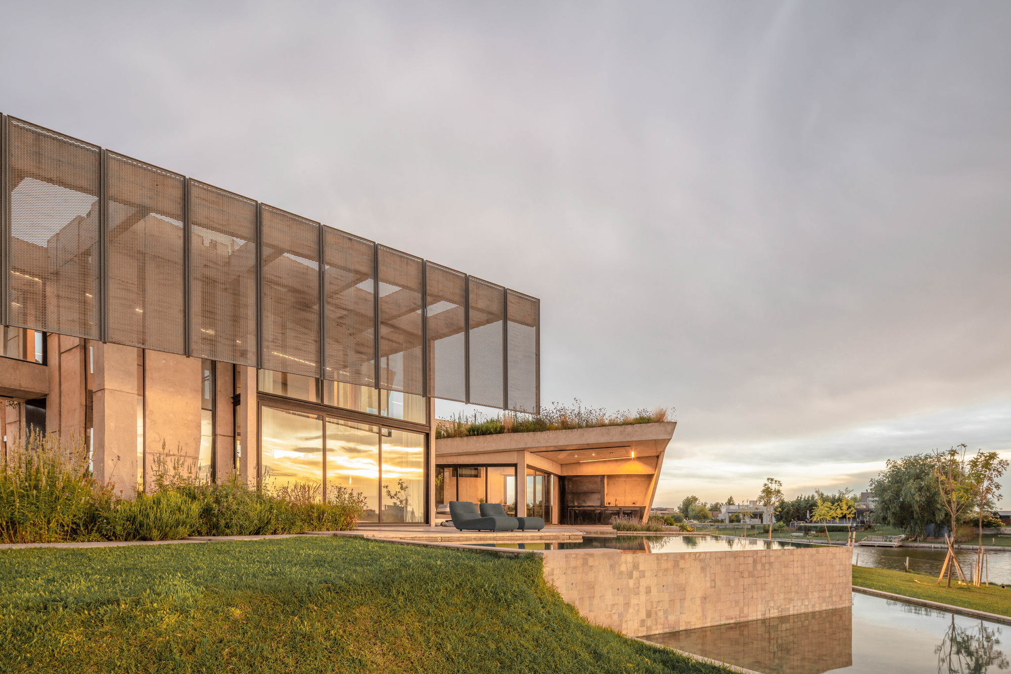 Perforated screen facade above a glazed ground floor and green roof, beside a reflecting pool in warm sunset light
