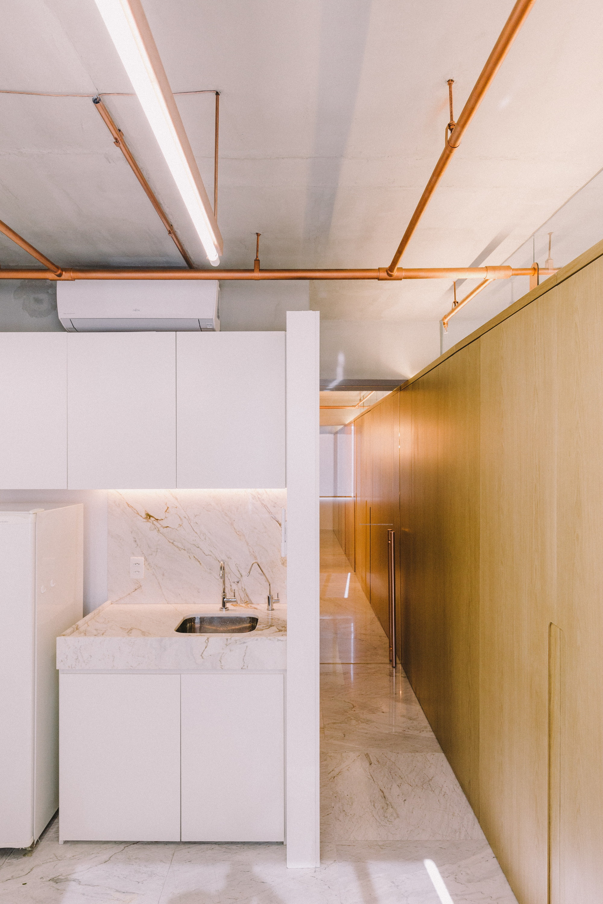 Narrow kitchenette with a marble splashback and sink beside a long oak wall of cabinet doors, copper pipes running overhead