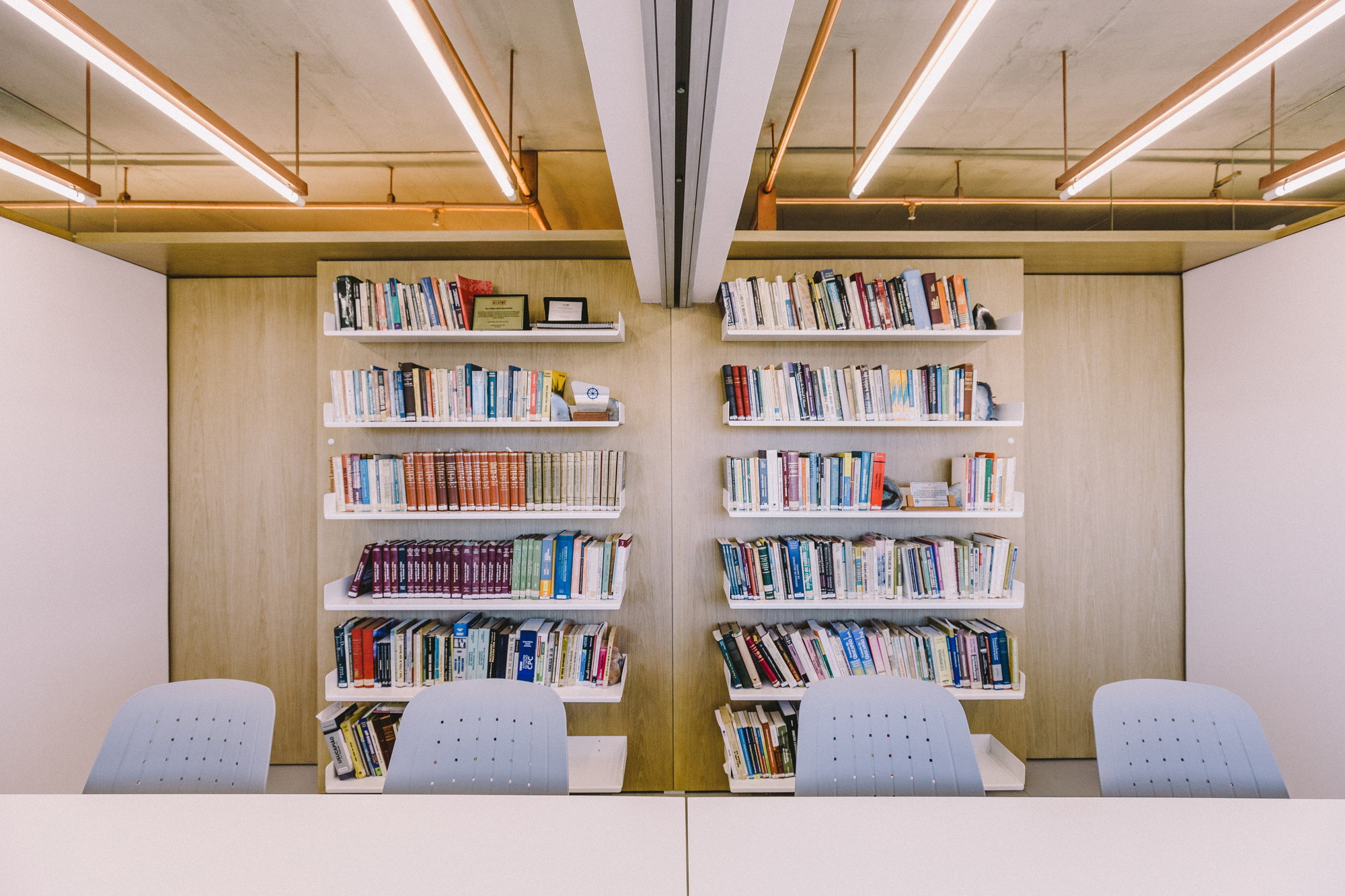 Wall of open shelves filled with books behind a shared white desk and four grey chairs, copper light tubes above