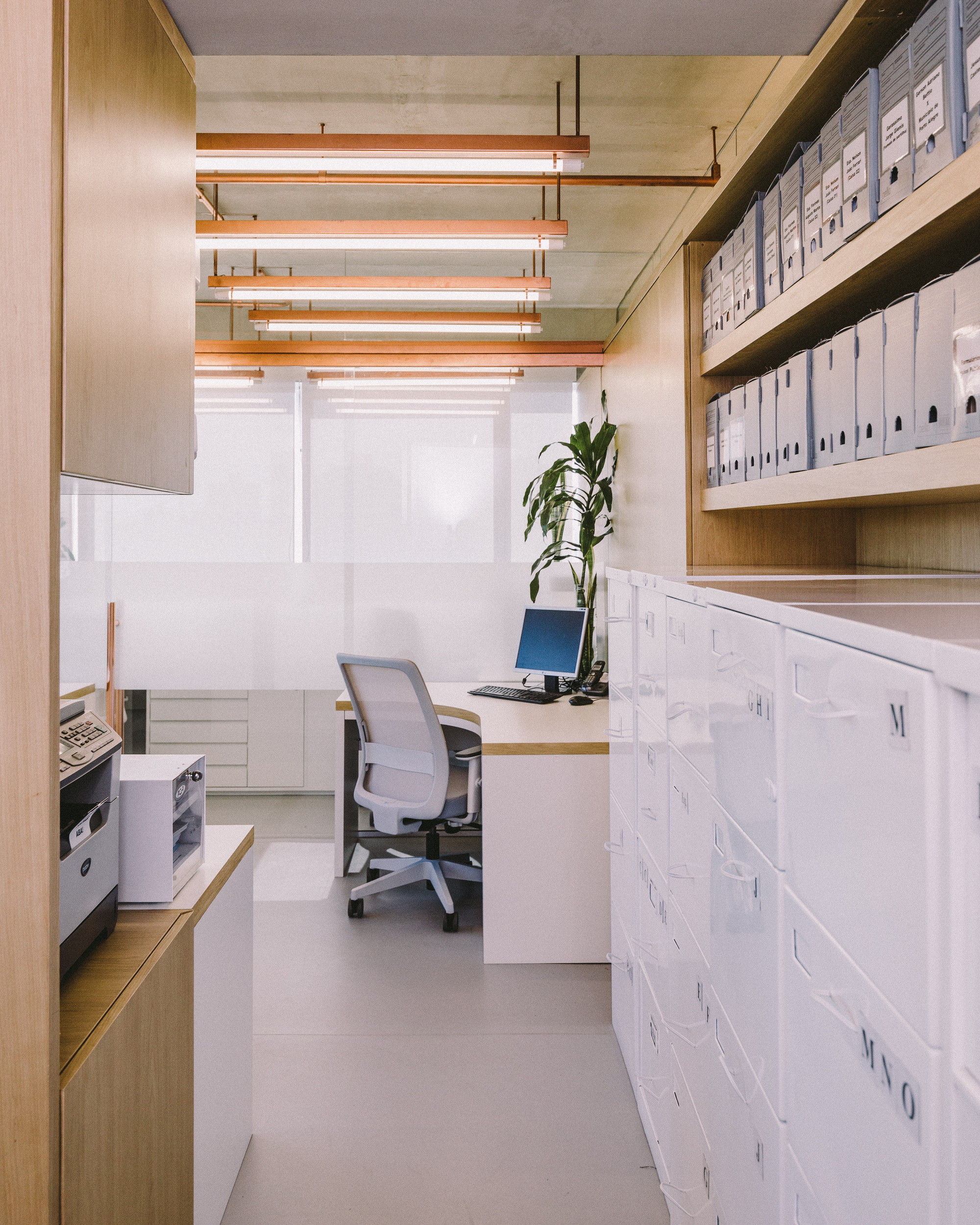 Narrow office aisle with white filing cabinets, archive boxes on oak shelves and copper-framed tube lights above a desk and plant
