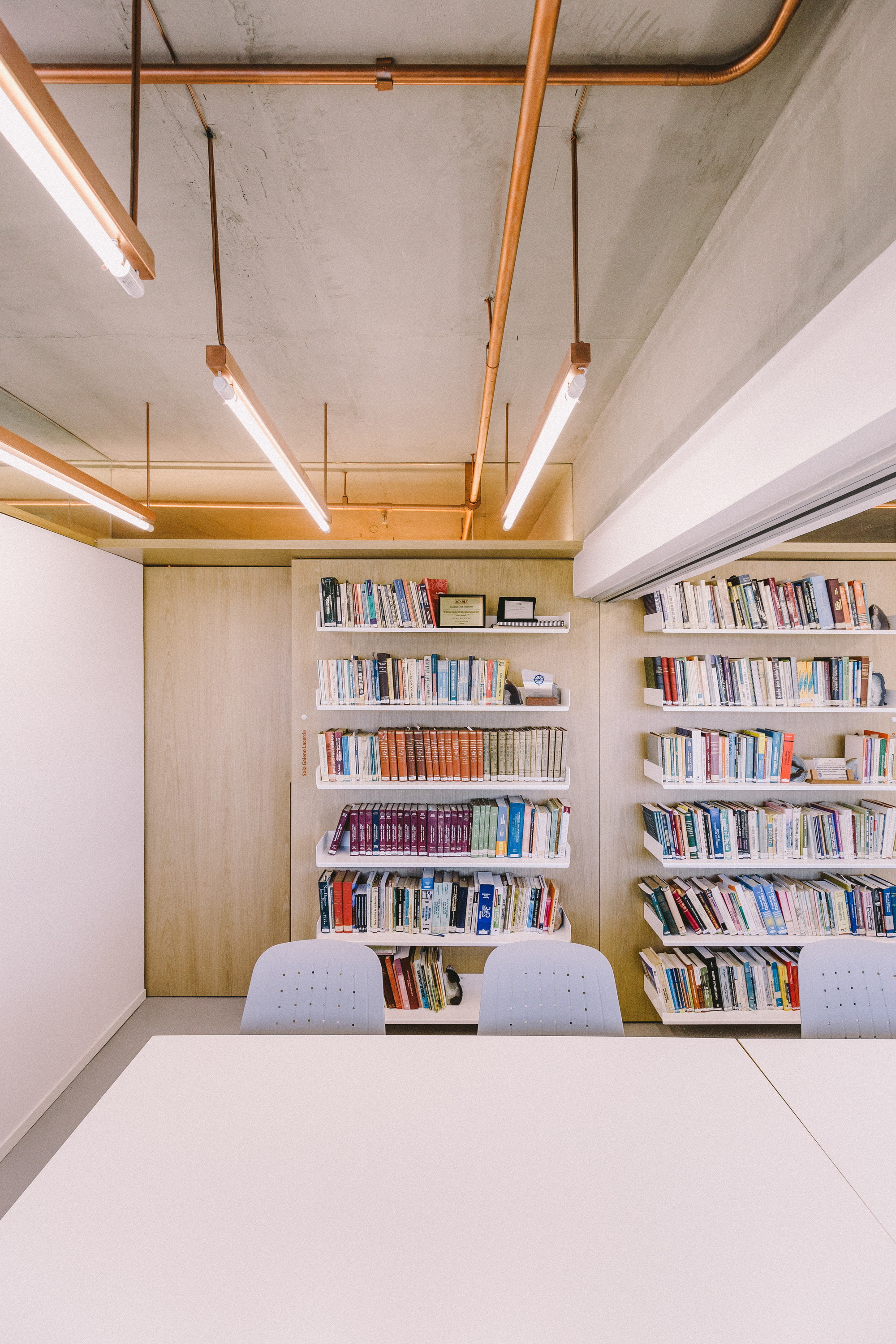 Meeting room with a white table facing wall shelves of books, under exposed concrete and copper pipes with tube lights