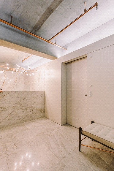 Marble-clad reception counter and flush white door beside a tufted bench, with copper pipes and a branching light overhead