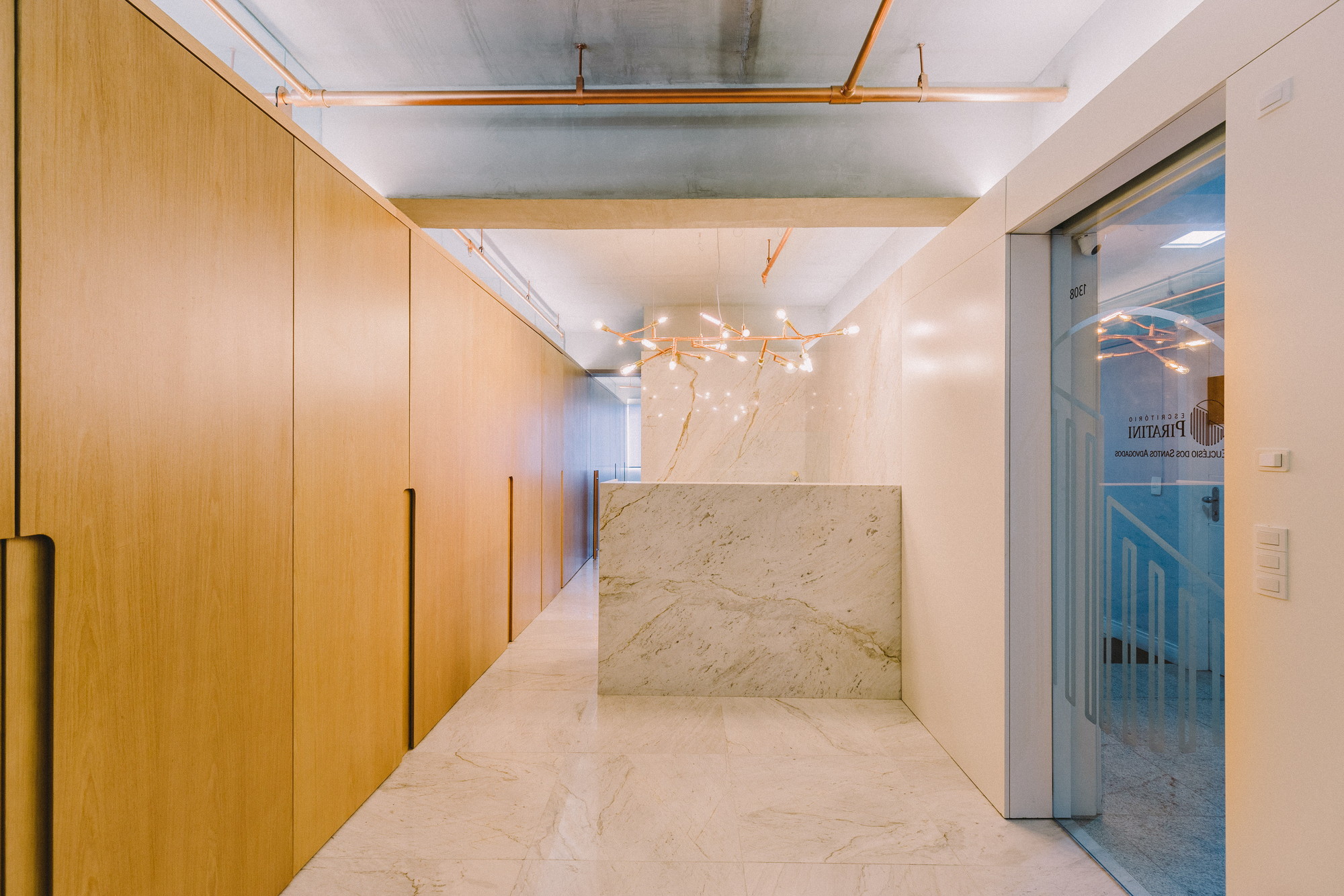 Corridor lined with oak cabinet doors leading to a marble reception block and glowing branch chandelier beside a glass door