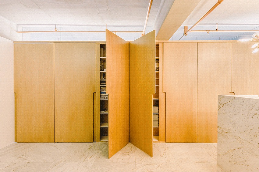 Pivoting oak veneer door swung open in a wall of tall cabinets, revealing shelves of books beside a marble counter edge