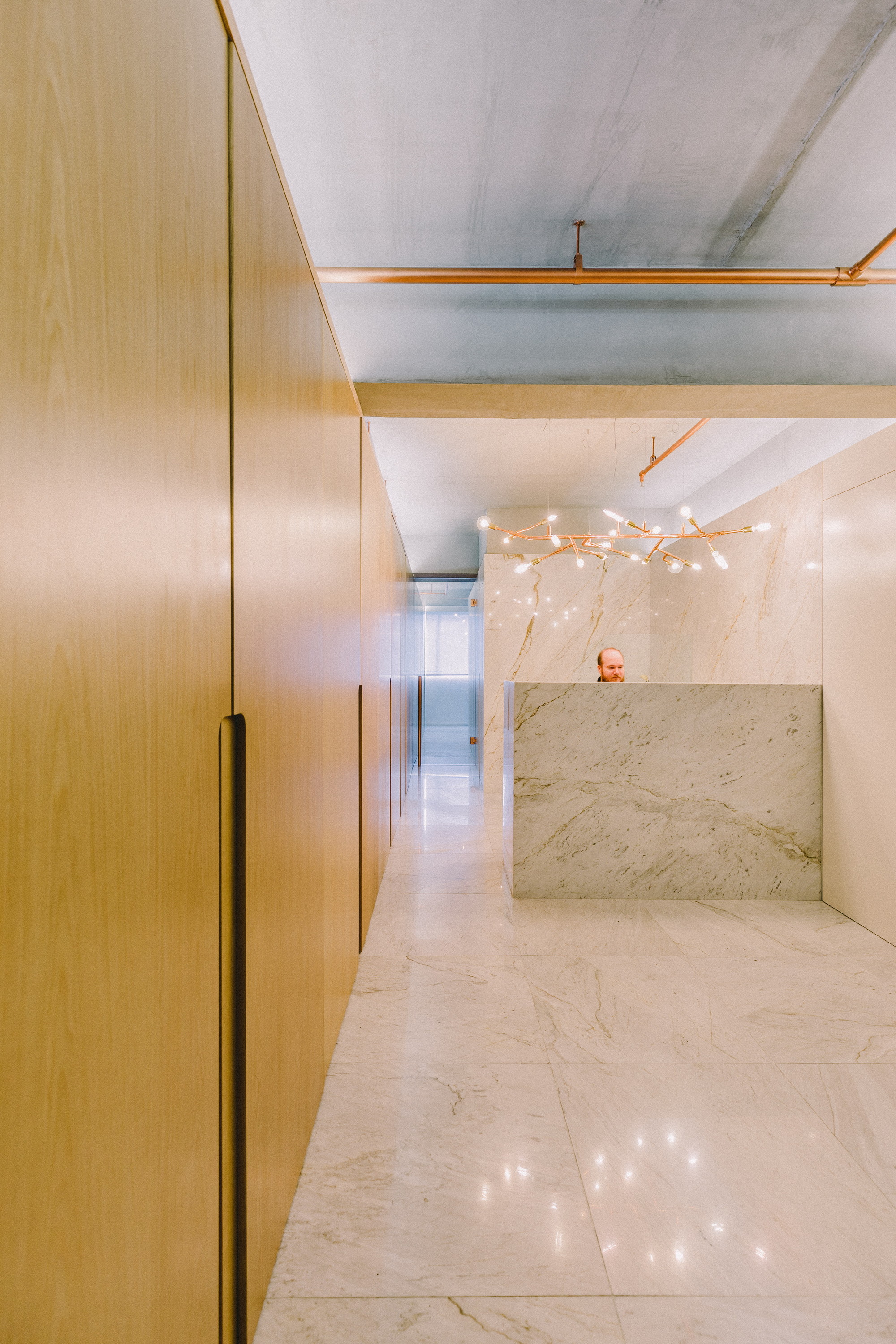 Corridor lined with oak cabinet fronts leading to a marble reception desk, a branching copper pendant light and a person behind it