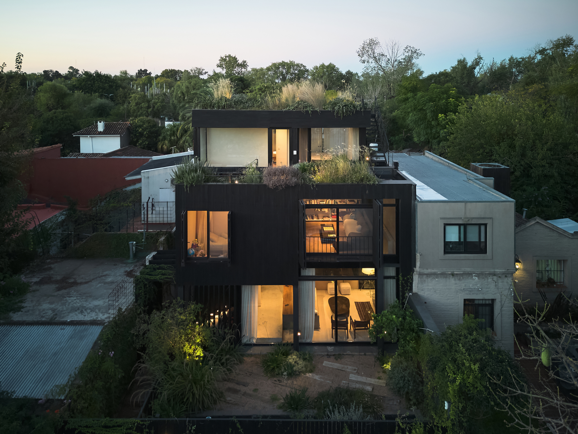 Rear facade of the dark timber volume with lit glazed rooms and a grassy roof garden at twilight