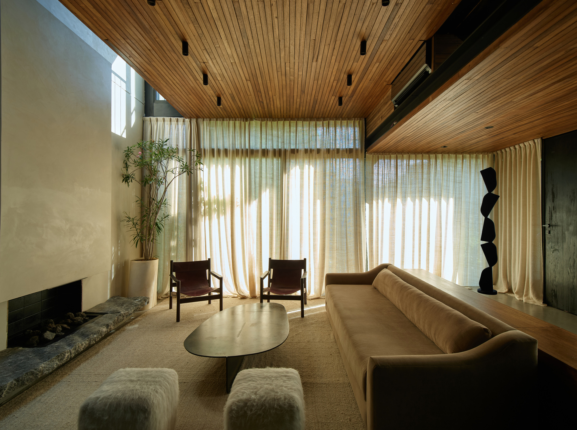 Living room with timber slat ceiling, sheer curtains glowing with sunlight, a camel sofa and a stone-edged fireplace