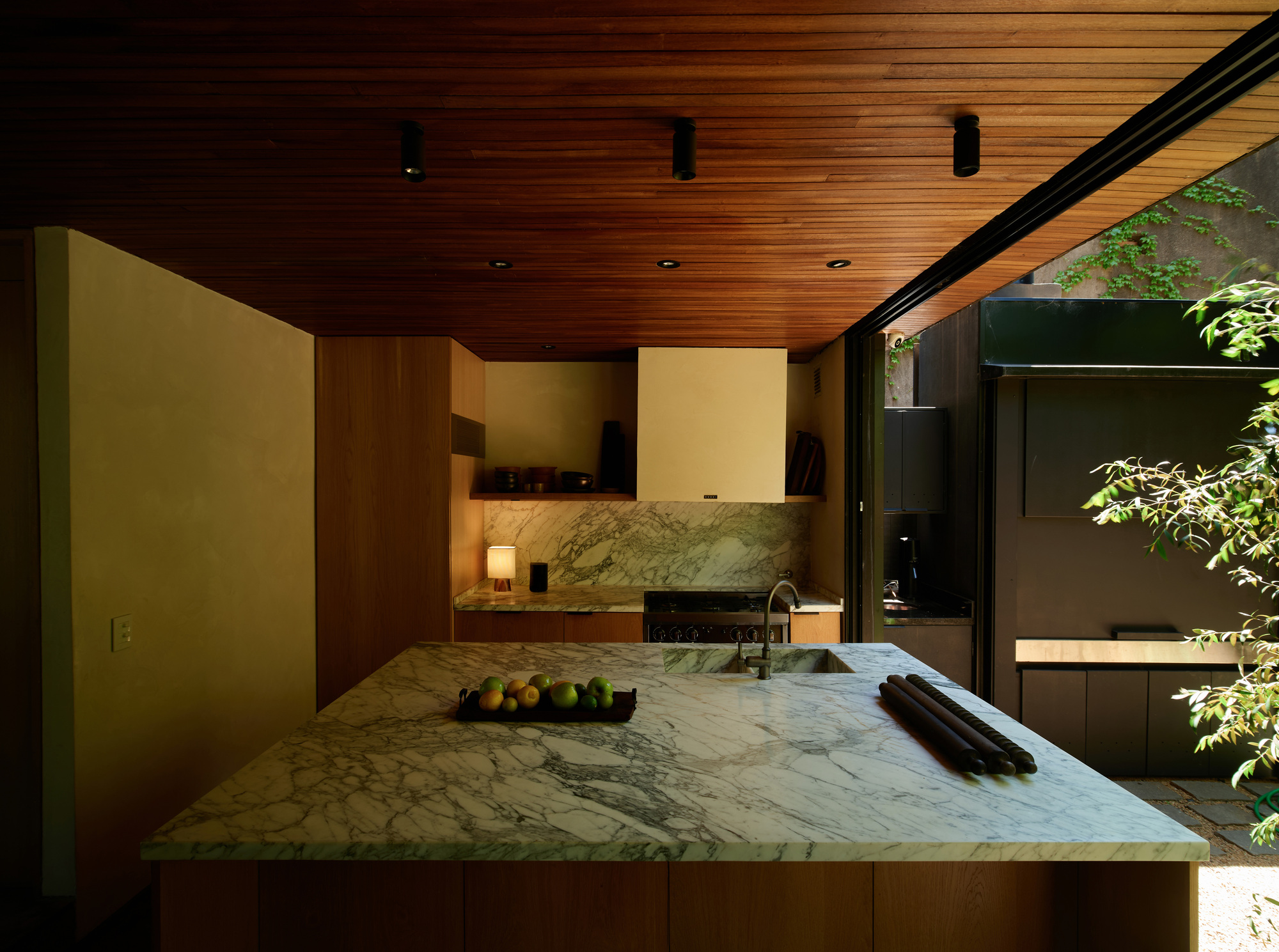 Kitchen island with veined marble top and fruit bowl beneath a timber ceiling, opening to a leafy courtyard