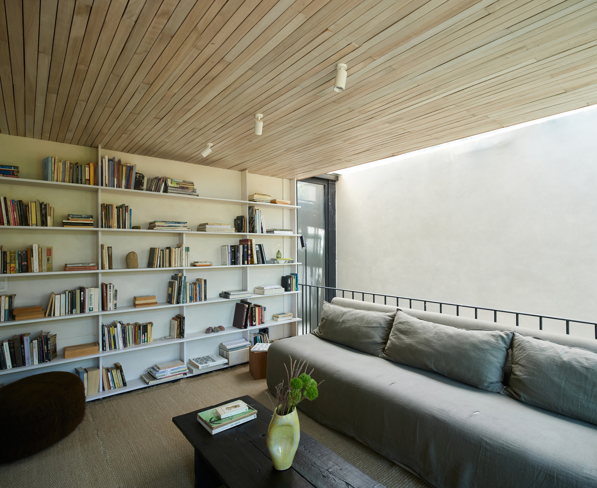 Reading corner with a full-wall white bookshelf, grey linen sofa and pale timber ceiling beside a bright light well