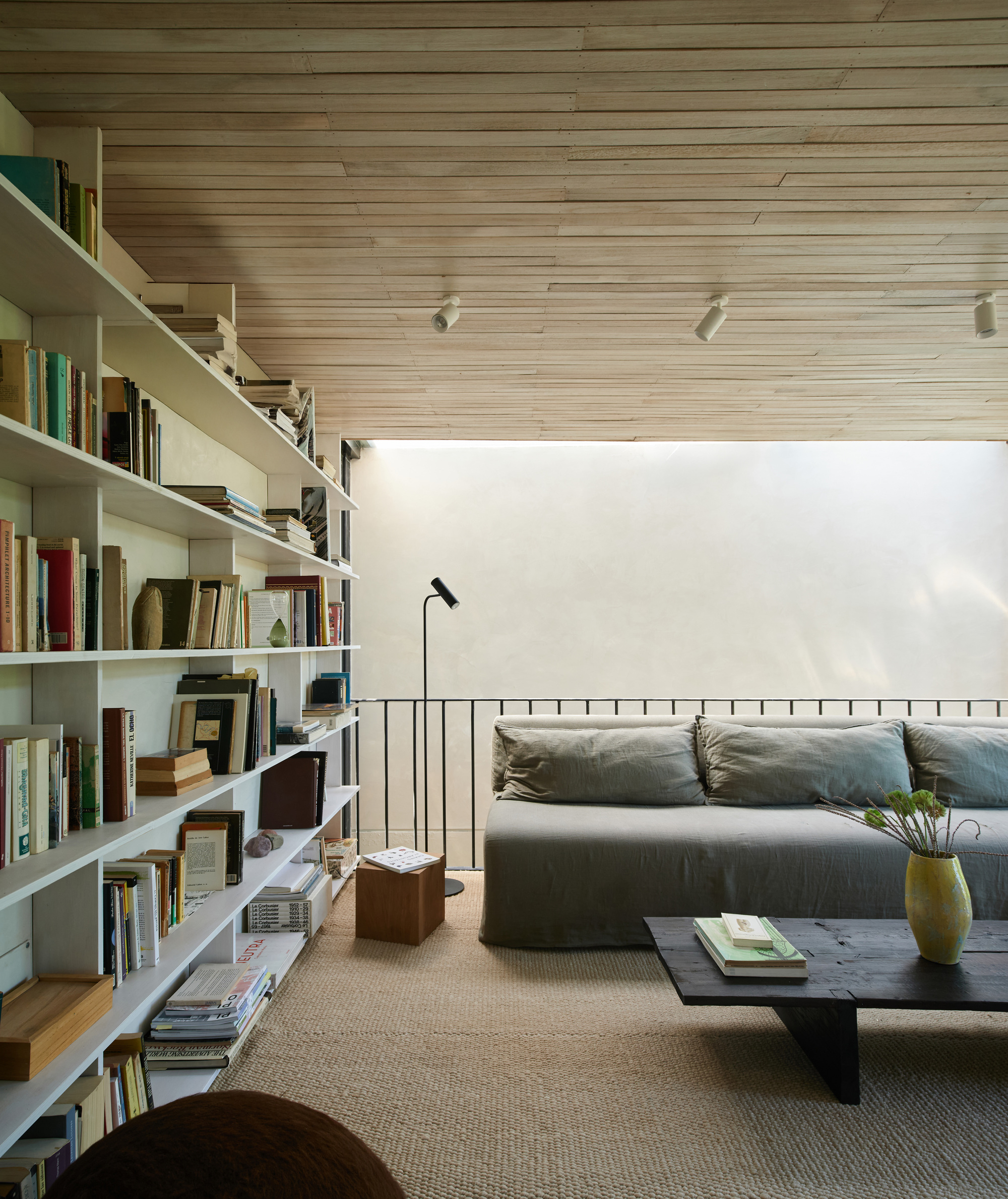 Living area with a built-in white bookshelf, grey linen sofa and low dark table beneath a pale timber plank ceiling in soft daylight