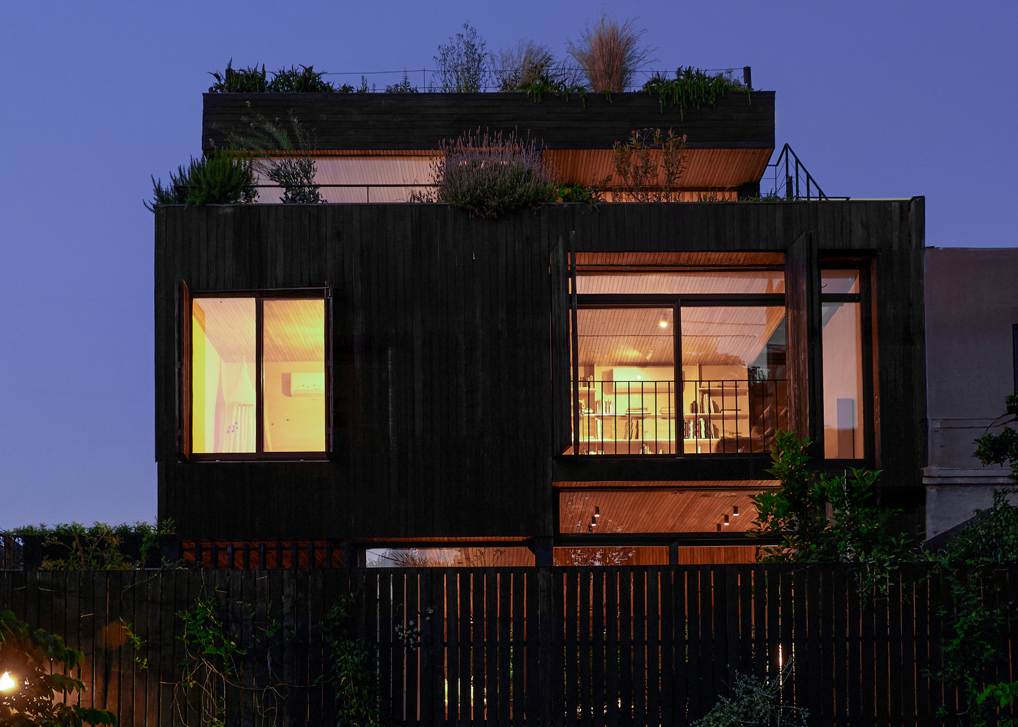 Dusk view of the charred timber house with lit windows and a planted rooftop terrace behind a slatted fence