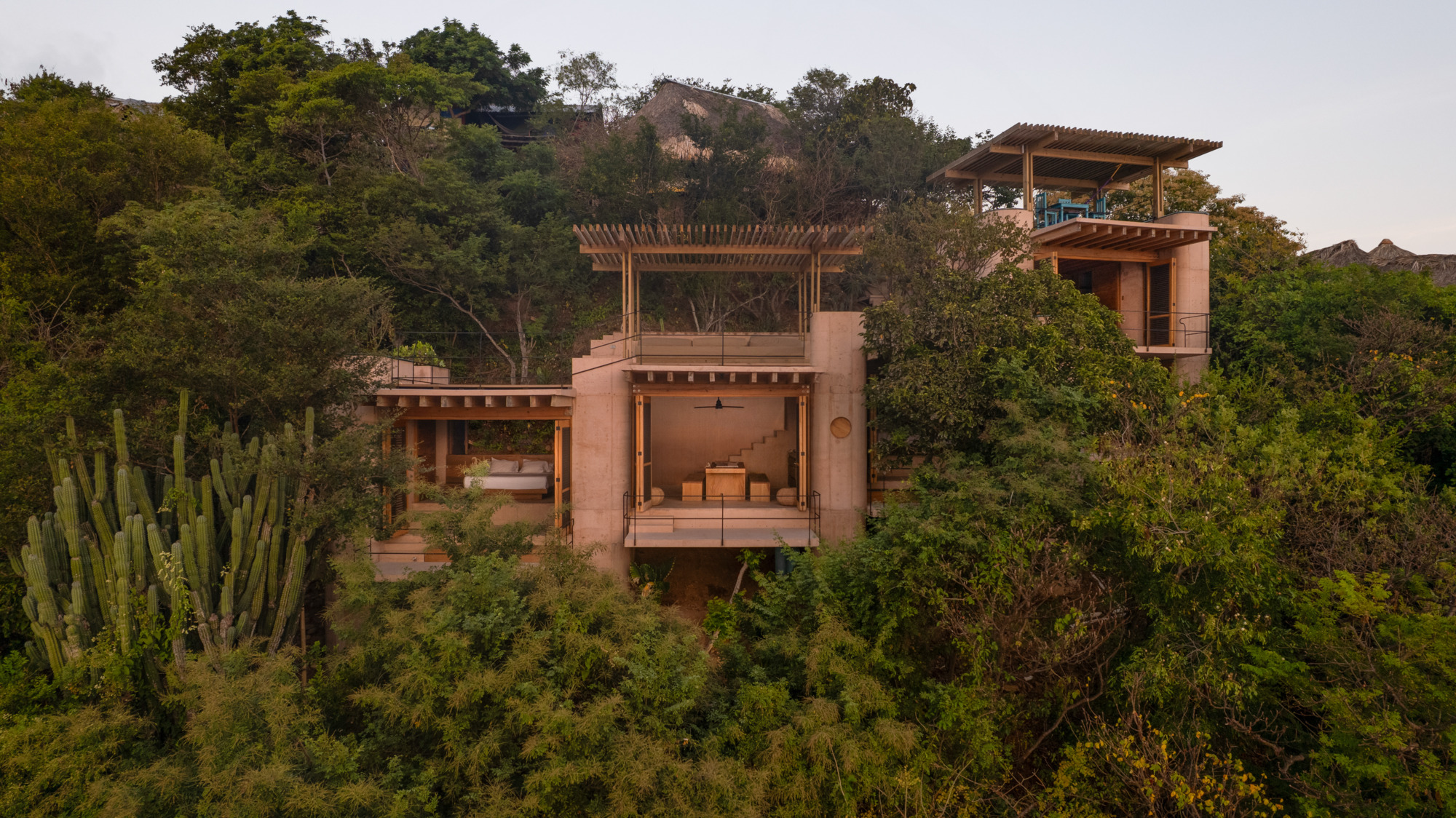 Drone view of stacked rammed earth volumes with timber pergola roofs rising through dense forest and tall cacti at dusk