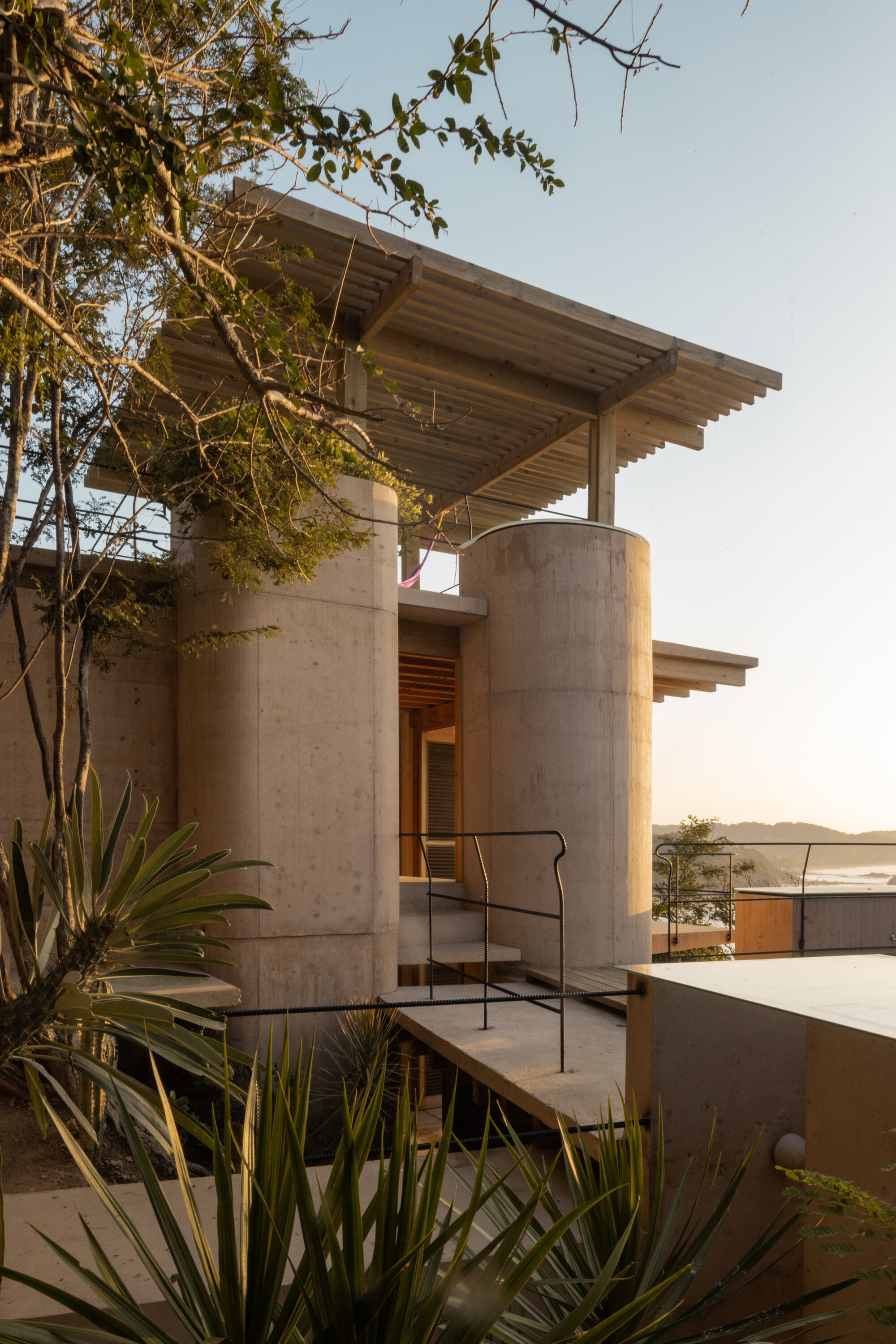 Two cylindrical concrete towers under a slatted timber roof, with agave leaves in the foreground and the sea glowing at sunset