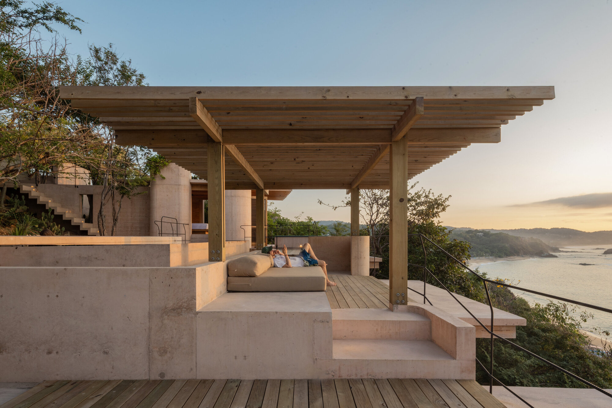 Timber pergola over a concrete daybed and wood deck, with a person reclining and a bay stretching out at sunset