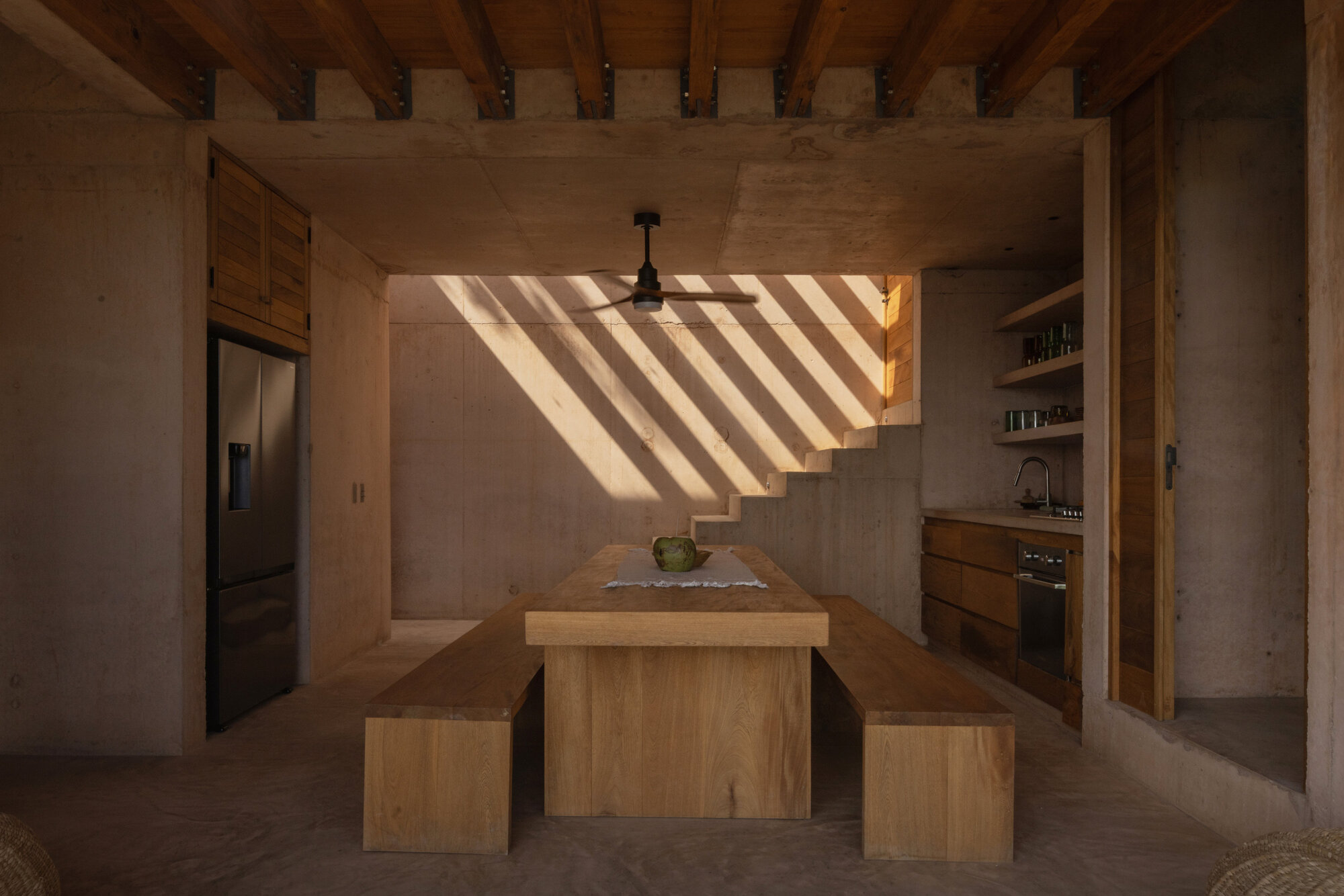 Open kitchen with a solid timber table and benches, where slatted sunlight falls across a concrete wall and stair