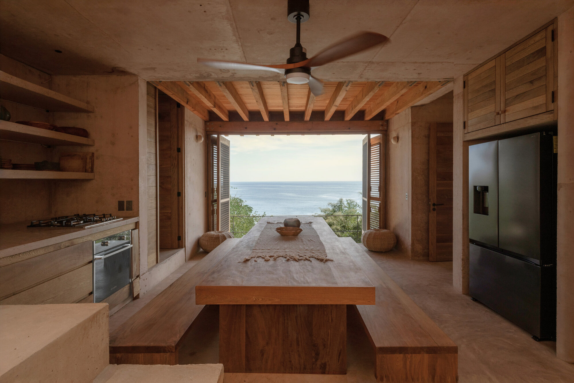 Concrete kitchen with a long timber table and benches facing an open wall toward the sea, daylight on the horizon