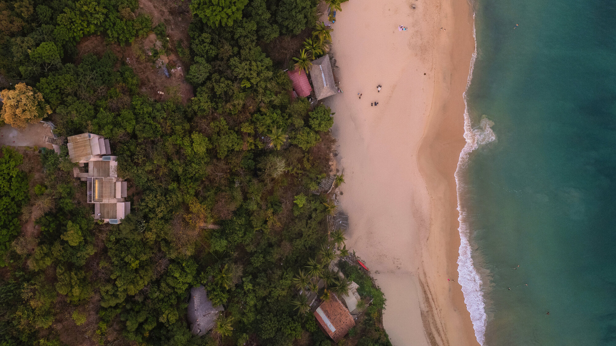 Drone view of timber-roofed pavilions among dense green forest beside a sandy beach and breaking surf at dusk