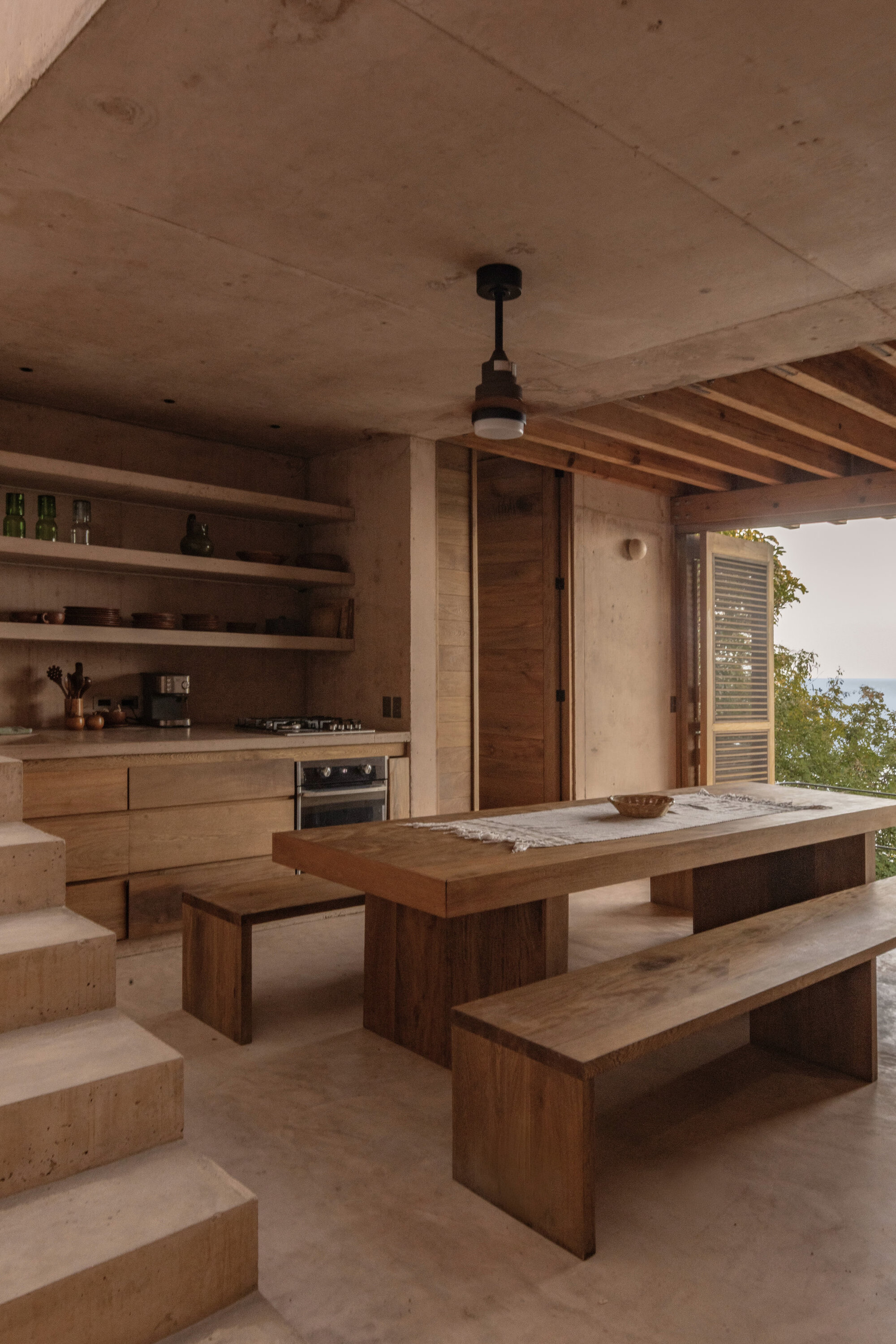 Open kitchen and dining area with concrete steps, wooden shelves and a plank table, ocean glimpsed through louvered shutters