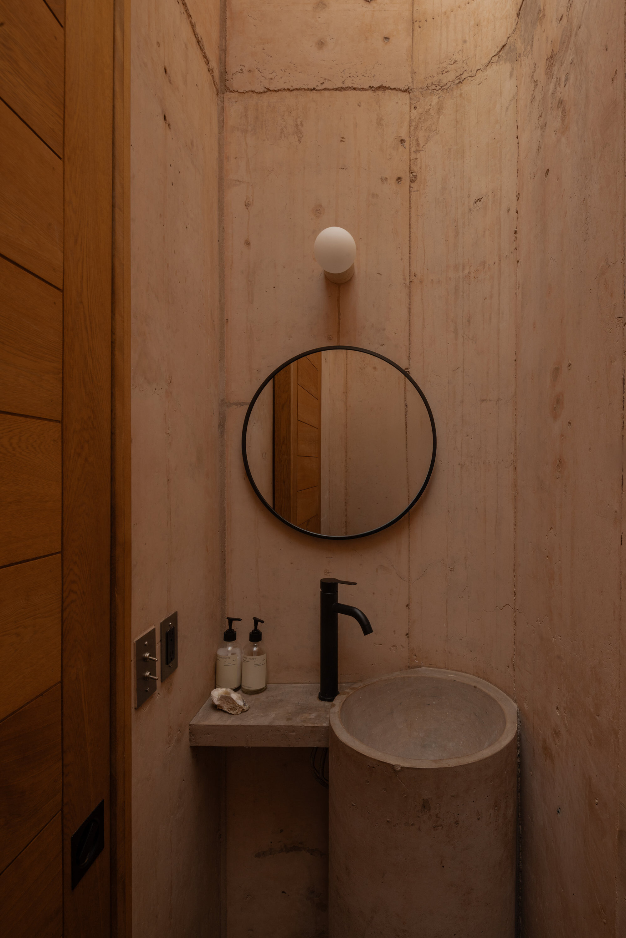 Small concrete washroom with a round black-framed mirror, cylindrical basin and black tap beside a timber door in soft light