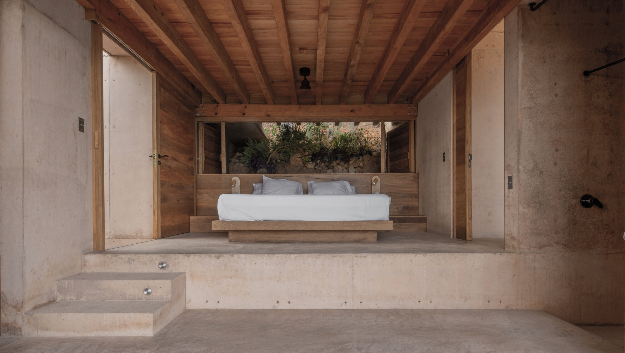 Raised concrete platform bedroom with a low timber bed, wood plank ceiling and a rock garden visible through the back opening