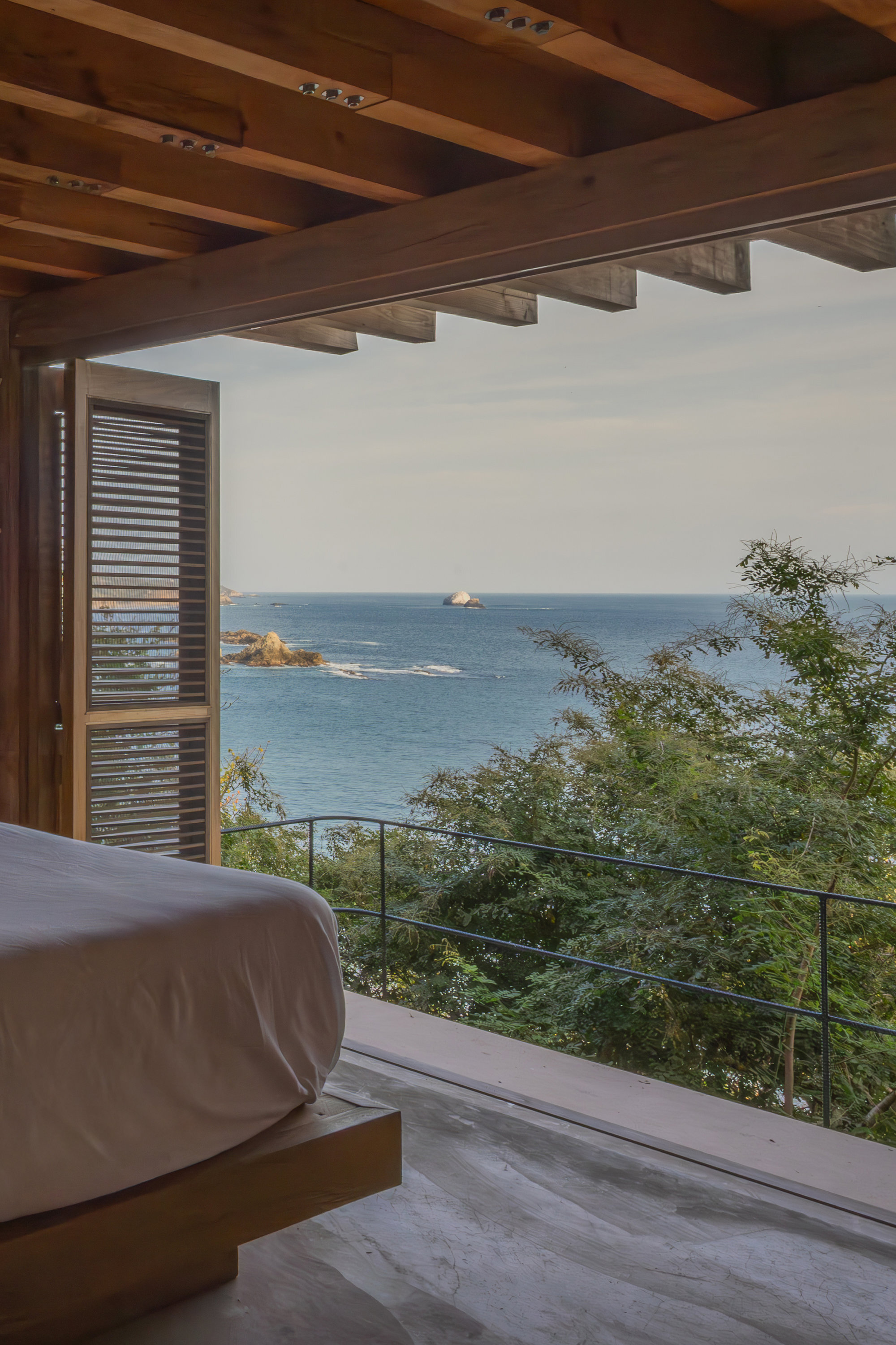 Bed beside an open timber-framed wall with a louvered shutter, looking past a thin railing to the sea in soft daylight