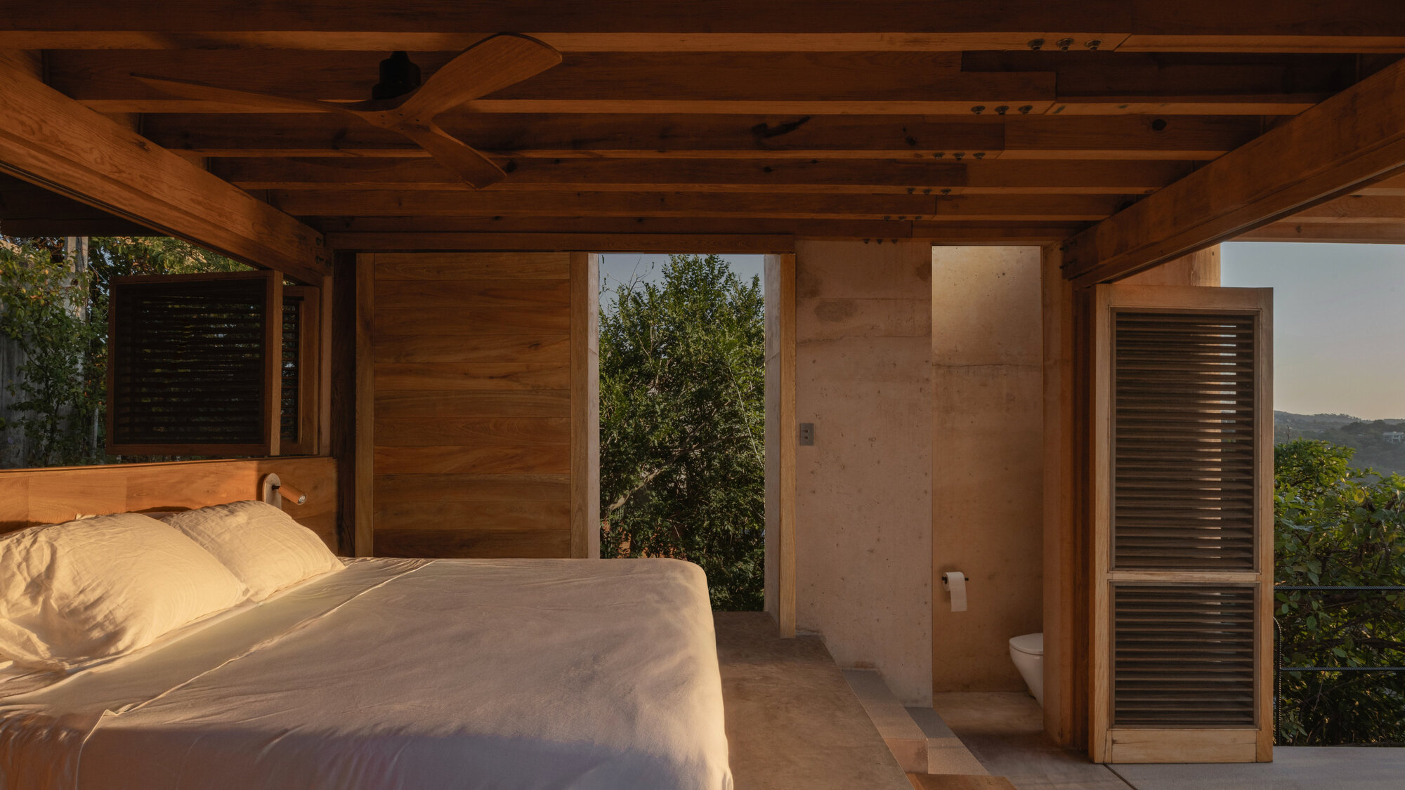 Open-sided bedroom under exposed timber beams with a ceiling fan, louvered shutter and concrete bathroom wall in warm evening light