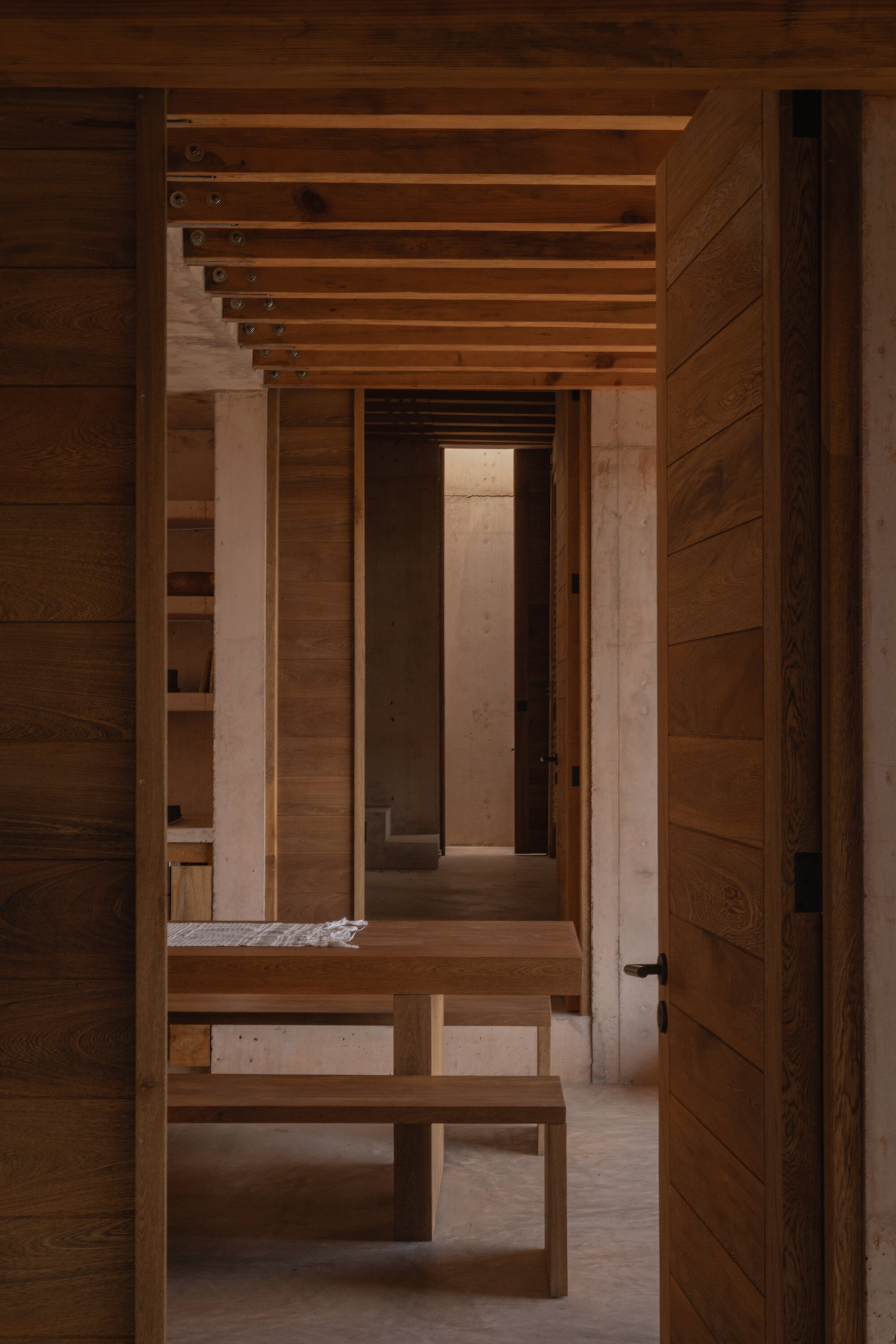 Enfilade view through timber-clad doorways toward a wooden table and bench, with exposed ceiling joists receding into the distance