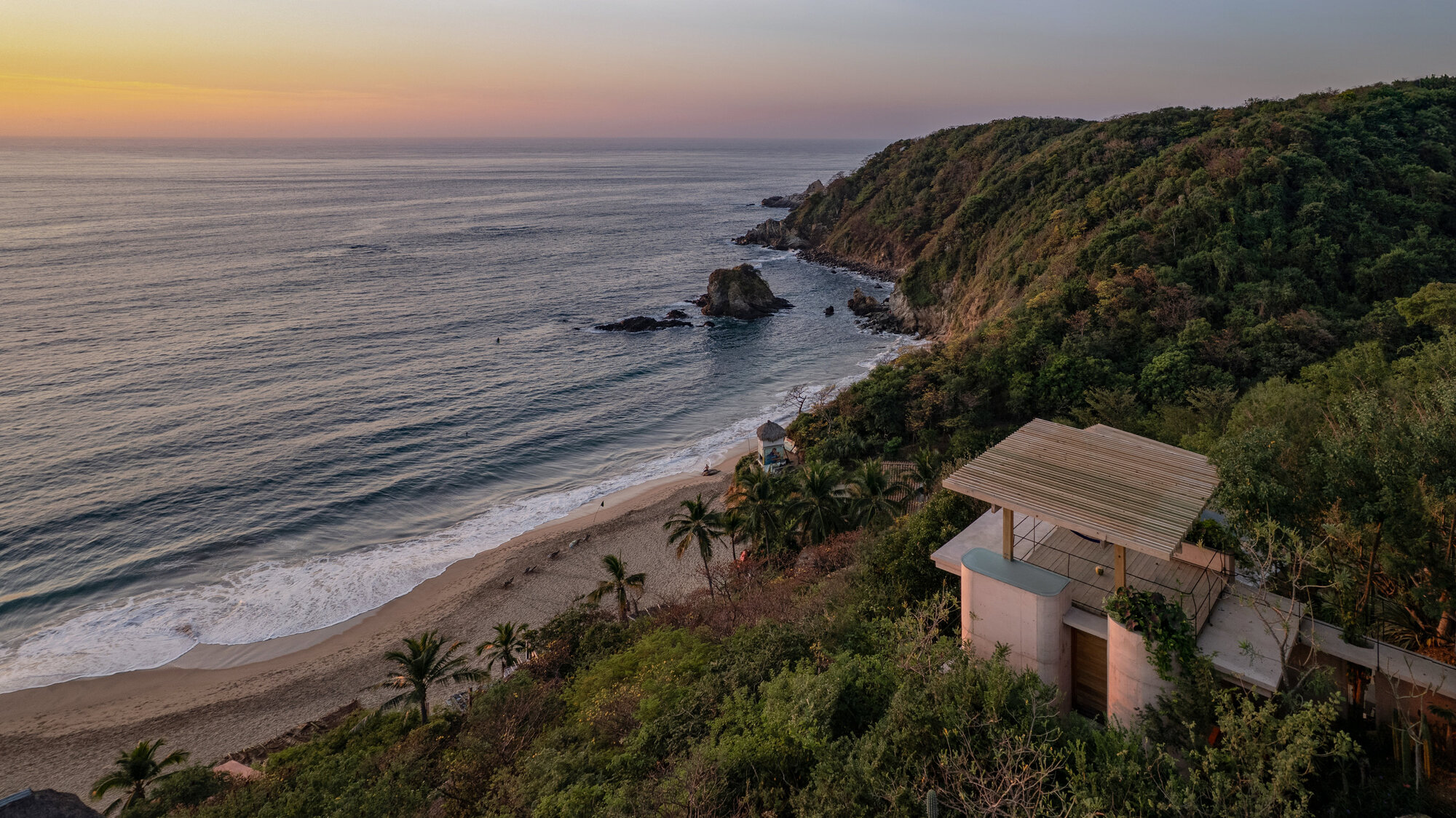 Aerial view of a rooftop pavilion with a slatted canopy on a forested headland above a sandy cove at sunset