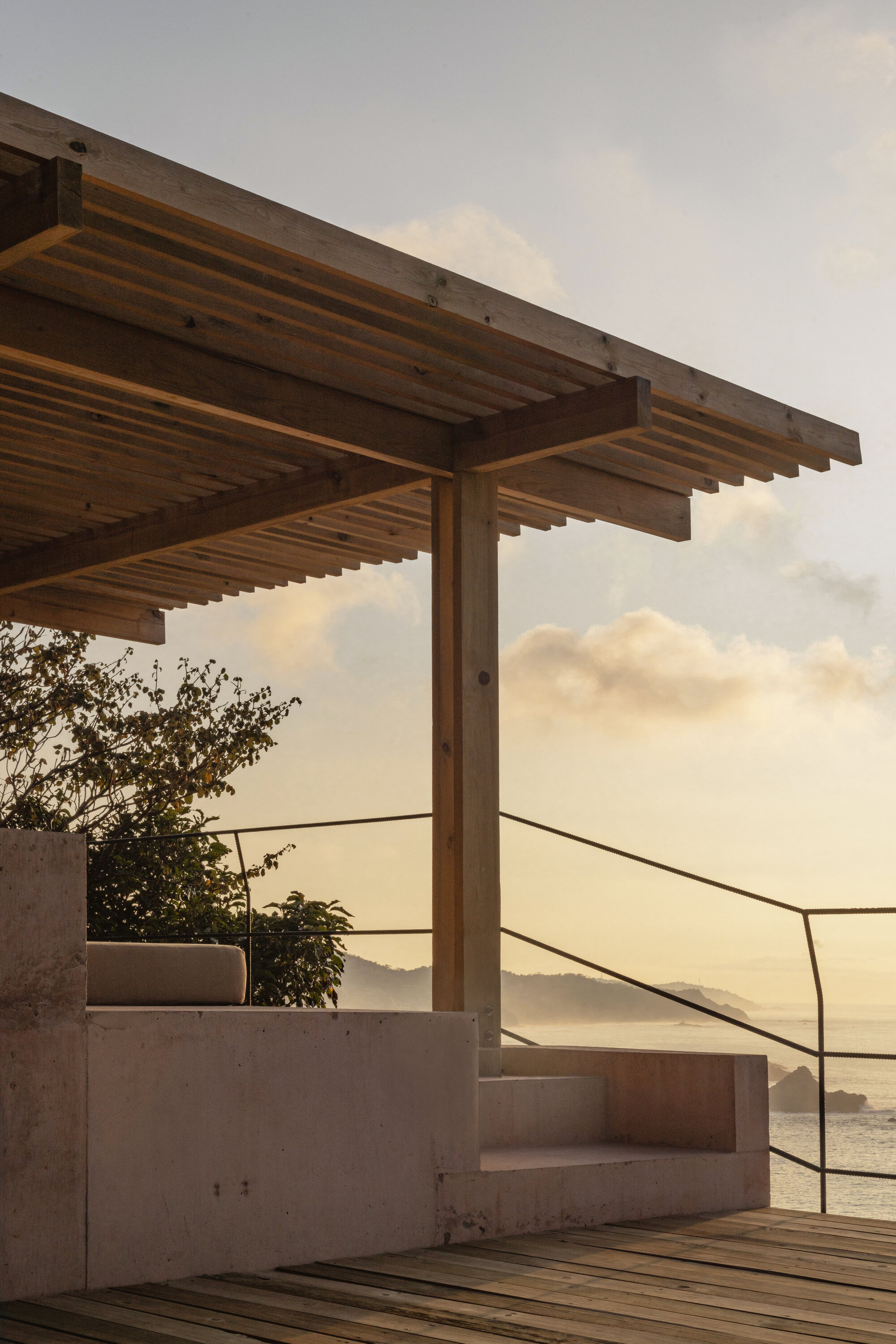 Timber pergola and concrete bench beside a wood deck, with a thin metal railing facing the ocean at sunset