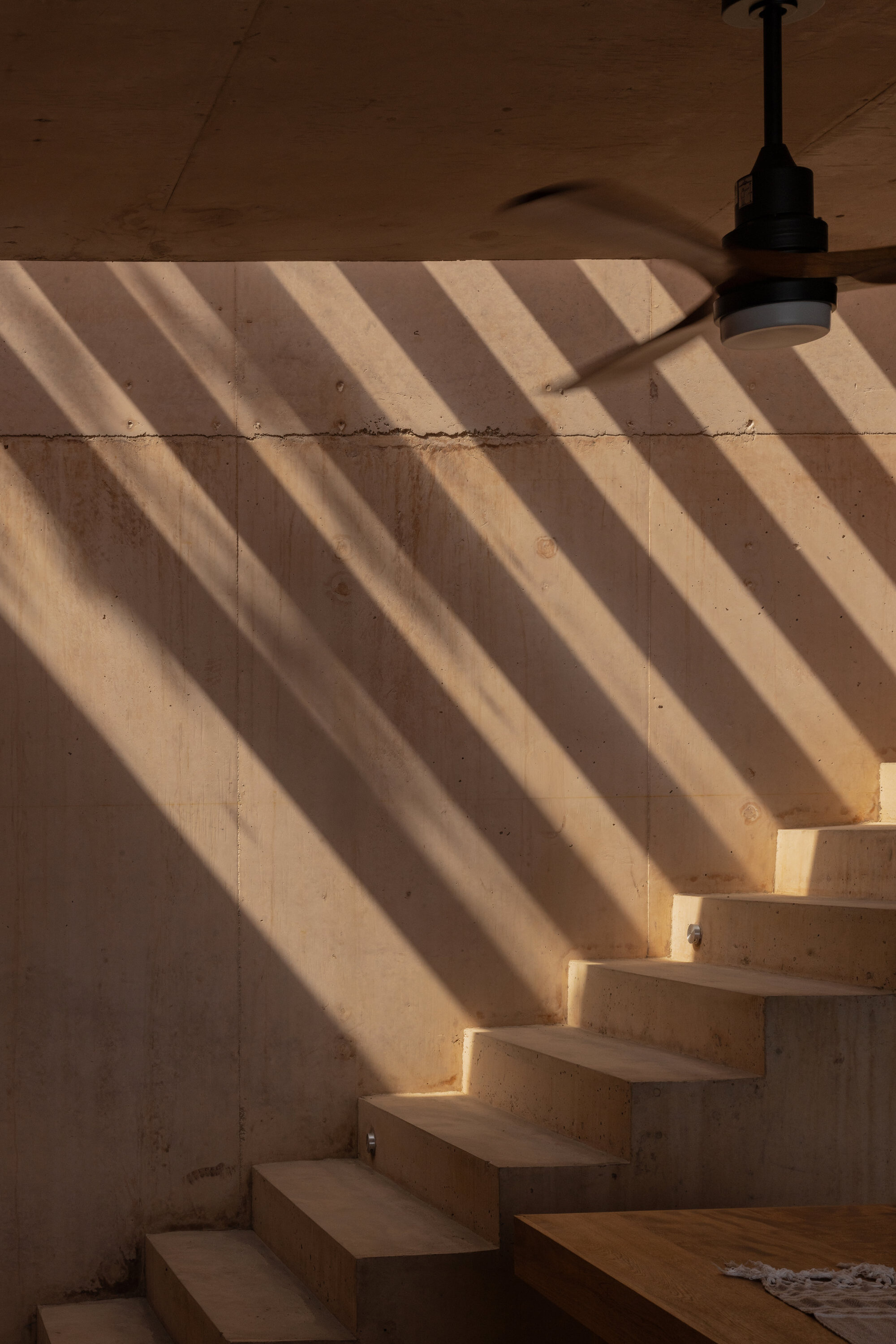 Concrete stair and wall striped with diagonal sunlight, with a ceiling fan blade blurred above a wooden platform